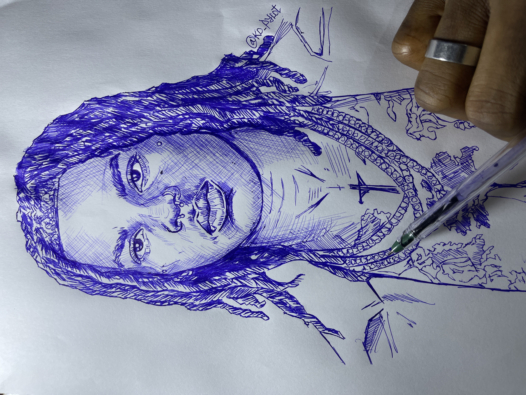 The image is a close-up, eye-level shot of a blue ink sketch on white paper, depicting the upper body and head of a person with dreadlocks. The sketch is detailed, with visible cross-hatching used to create shading and texture, particularly on the hair and face. The subject is wearing multiple large chain necklaces and a pendant shaped like a cross. The lower right portion of the image shows a person's hand holding a blue ink pen, with a silver ring on one finger, suggesting the sketch is in progress. There are subtle tattoo-like designs visible on the subject's chest area in the sketch. The background is plain white paper. The image does not contain any elements that would indicate its location in Jos, Nigeria.