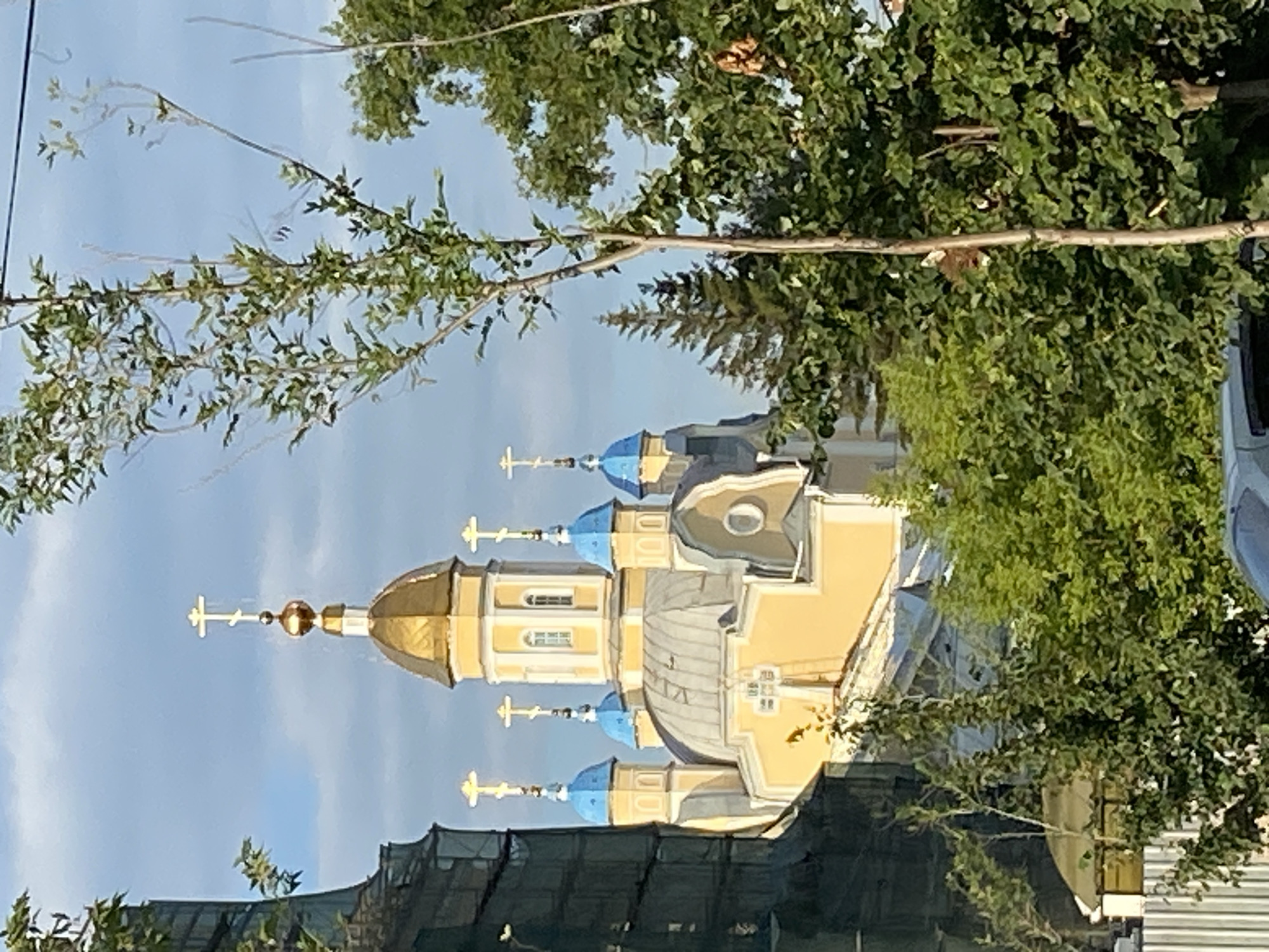 The image depicts an outdoor scene featuring a church with multiple domes, some gilded and others a vibrant blue. The church's facade is painted a light yellow, and it has several windows. Crosses adorn the tops of the domes. The building is partially obscured by lush green foliage from trees in the foreground. There is some scaffolding or construction netting visible at the base of the church. The sky is a light blue, suggesting it is daytime. The overall scene appears serene and possibly devotional, with the sunlit church and greenery creating a pleasant atmosphere. The image's orientation is sideways.