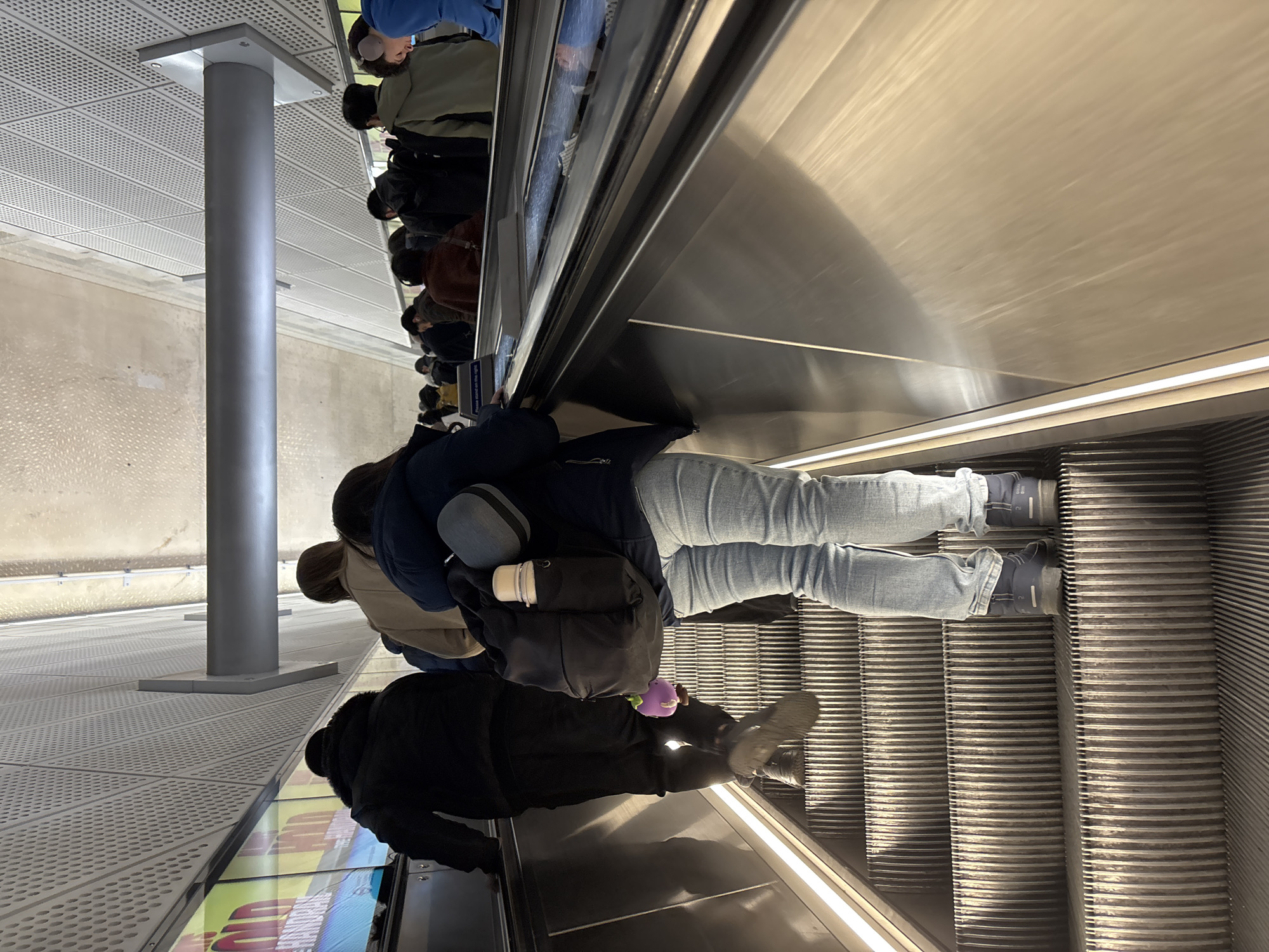 A medium shot shows an escalator ascending upwards. A line of people is taking the escalator. The escalator handrail is metallic and shiny. The escalator steps are metal and ridged. The people wear casual winter clothing. A metal support pole is visible to the left of the escalator, running from the ground up to a metallic perforated ceiling. The ground shows patterns of squares and rectangles, some of which have illuminated displays. The location is likely a station within the city of London, United Kingdom.