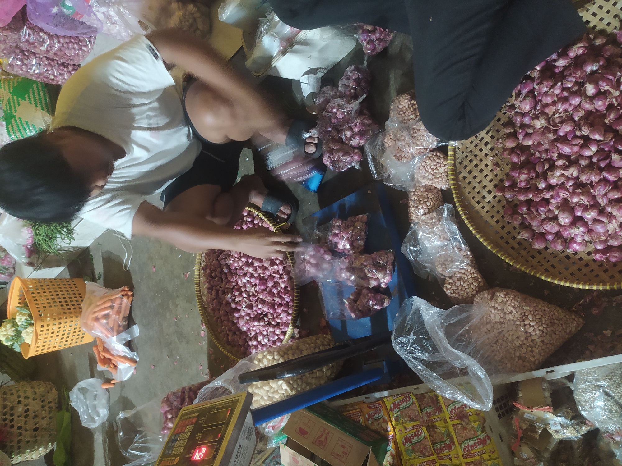 A person is crouching down on a concrete floor in a market stall in Kupang, Indonesia, sorting through shallots. The person is wearing a white t-shirt and black shorts. In front of the person, there is a large woven basket filled with shallots. To the left of the basket, there is a digital scale and a box of what appear to be packaged goods. Behind the shallots, there are several plastic bags filled with peanuts and more shallots. In the background, there are more market goods, including carrots in plastic bags and a basket of cauliflower. The lighting suggests it is either nighttime or an indoor market.