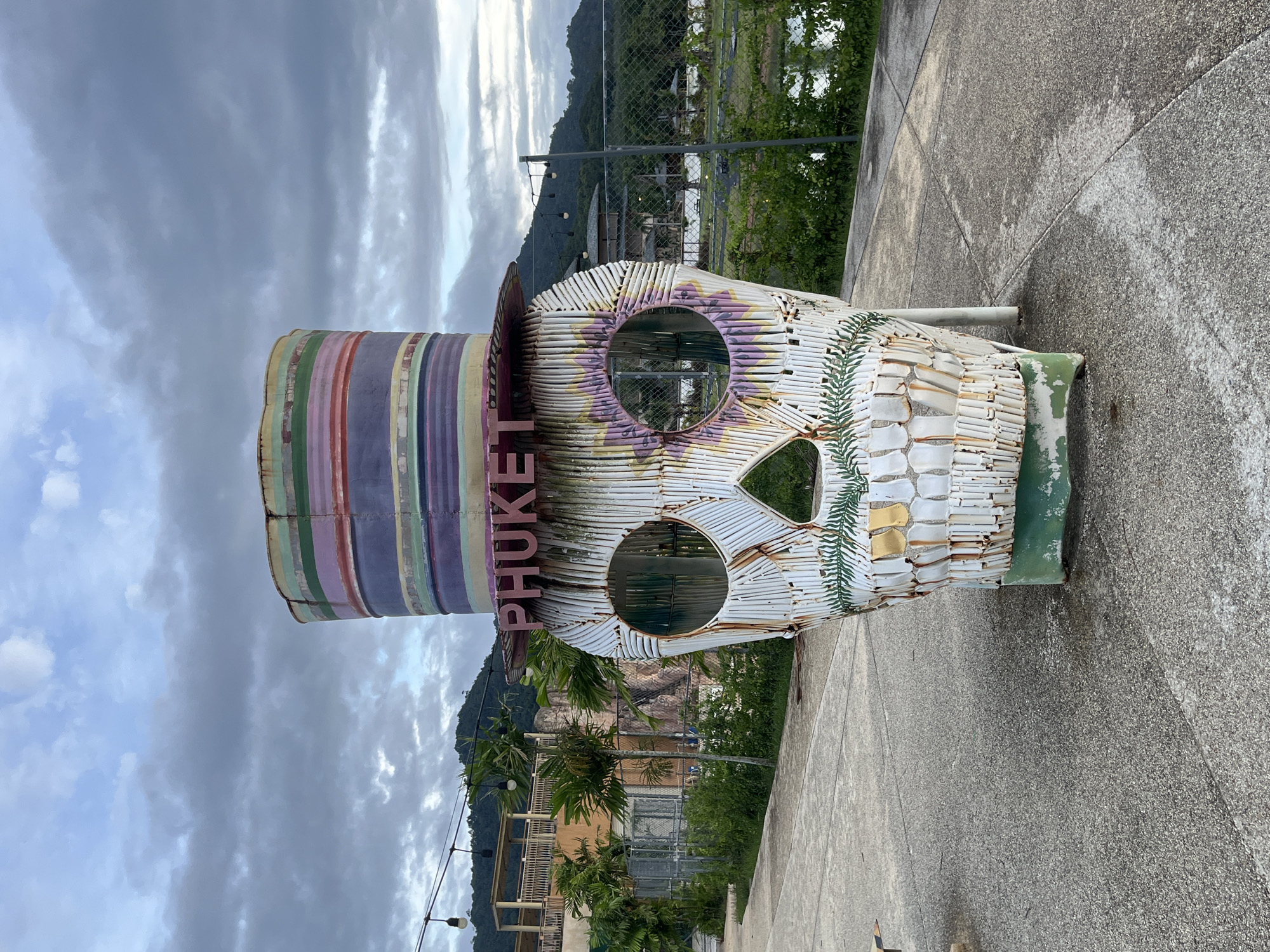This image, taken in Choeng Thale, Thailand, depicts an outdoor scene under an overcast sky. A large, stylized skull sculpture with a top hat occupies the central foreground. The sculpture is constructed from corrugated metal panels, showing signs of weathering, rust, and chipped paint. The skull portion is primarily white, featuring large circular eye sockets, a triangular nose opening, and a suggested mouth. Green fern-like patterns are painted below the nose, while purple and yellow floral patterns adorn the areas around the eye sockets. The base of the skull is painted green.
The top hat is cylindrical with a wide brim, also made of weathered, painted metal. It displays horizontal stripes in various colors, including purple, green, light blue, pink, red, yellow, dark green, orange, light purple, brown, and light pink. The word "PHUKET" is spelled in raised pink letters on the front brim of the hat.
The sculpture rests on a light grey concrete or paved surface that exhibits wear and discoloration. In the midground, a series of bare light bulbs are strung overhead. To the left of the sculpture, several small palm trees and green bushes are visible, beyond which stands a multi-story building with open-air sections and railings. A chain-link fence runs across the midground, separating the immediate area from the background. To the right, behind the fence, is a body of water bordered by green vegetation and distant structures. The background features rolling hills or mountains densely covered in trees, all beneath a sky filled with grey and white clouds. No individuals are present in the scene.