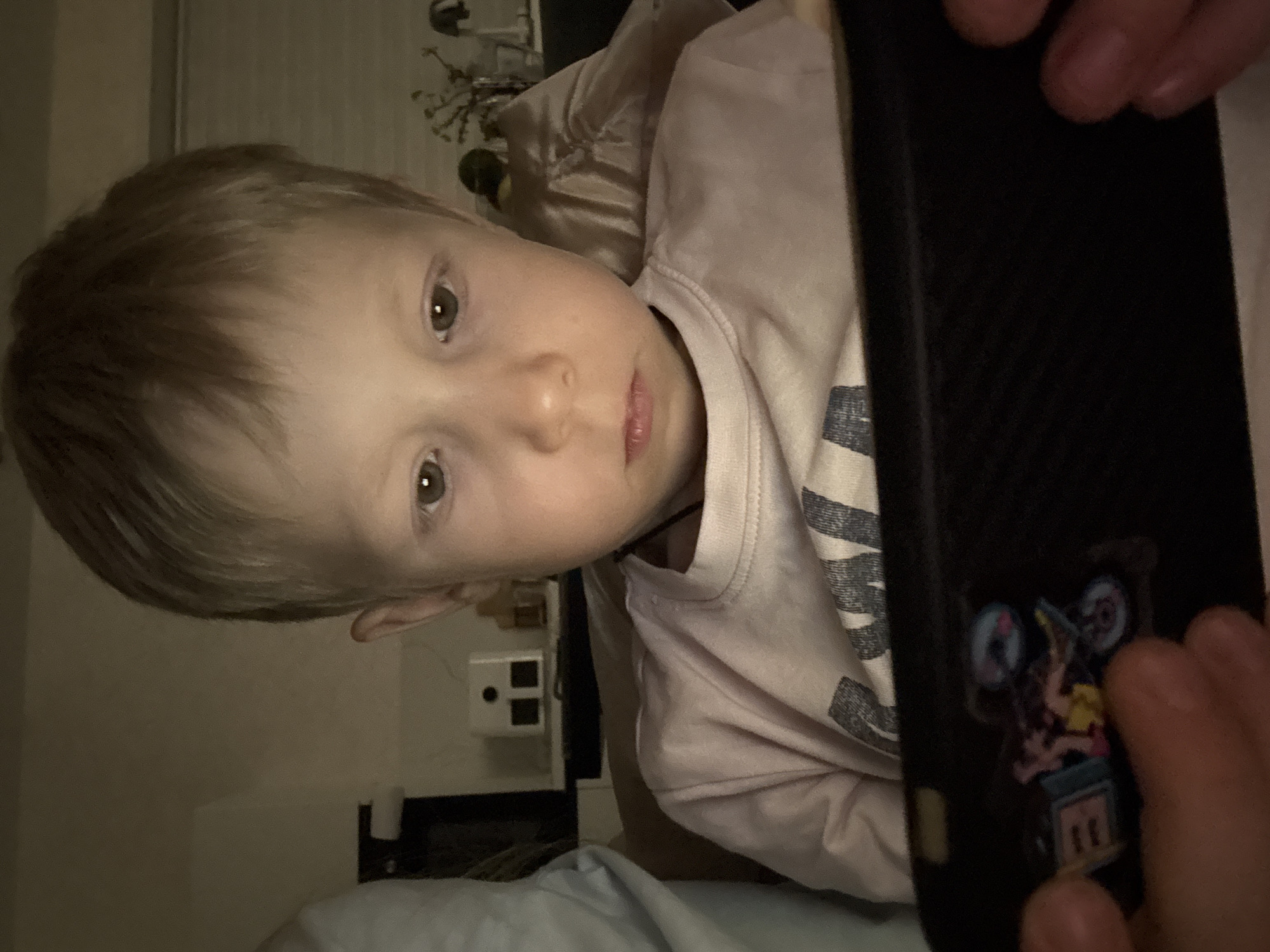 A blonde-haired child with light skin is lying down and looking at a mobile phone. The phone is held in the hands of an adult whose fingers are visible on the edges of the phone. The child is wearing a light pink long-sleeved shirt with a grey design on it. The background shows a room with a window covered by blinds and a small bedside table with objects on it. The location is unknown and cannot be determined from this image. The city of Chaikovsky, Russia, cannot be confirmed.