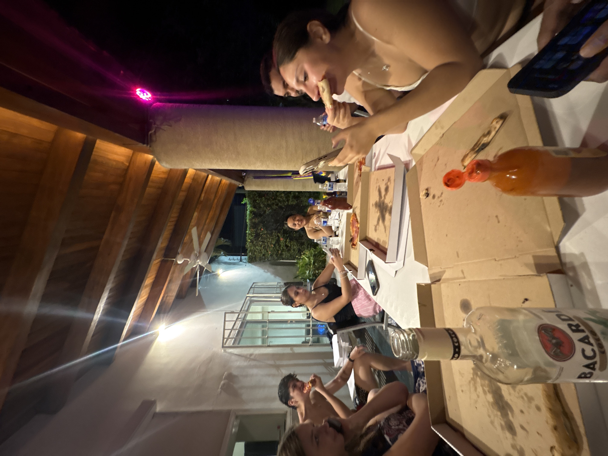 A group of people is gathered around a table on a patio at night, eating pizza and drinking. The patio is under a wooden roof with pink decorative lighting. Several pizza boxes are on the table, along with a bottle of Bacardi rum and a bottle of hot sauce. The patio appears to be connected to a house with a fan on the porch. The location is most likely in a residential area in Puerto Escondido, Mexico.