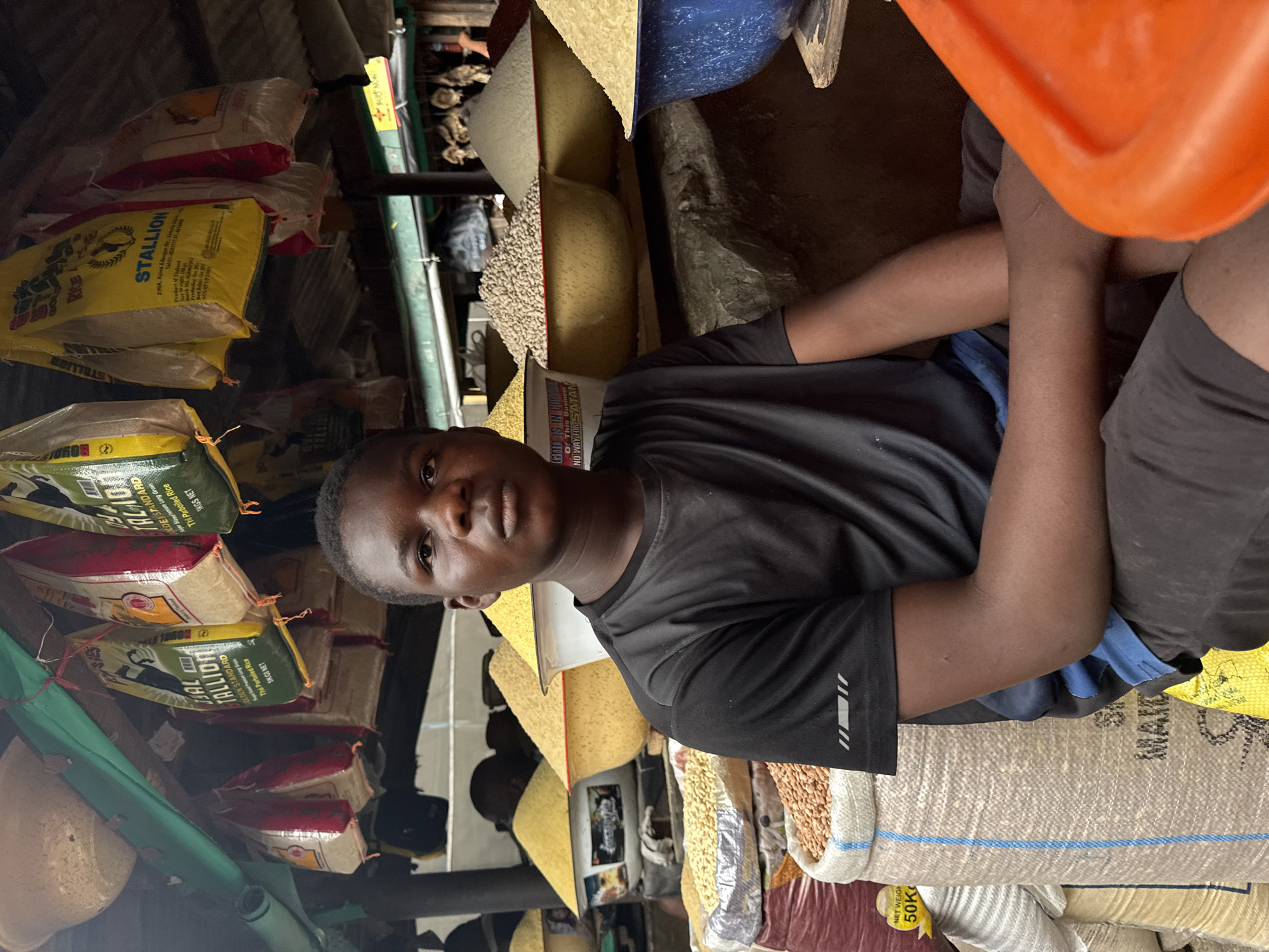 The image depicts a young Black male, appearing to be in his late teens, sitting in what appears to be a food market stall in Lagos, Nigeria. He is wearing a black t-shirt with a subtle grey stripe design on the sleeve and dark shorts with a blue waistband. His gaze is directed towards the camera.
The stall is filled with various sacks of grains and legumes. Behind him, large, colorful bags, some labeled "STALLION GOLDEN STANDARD PARBOILED RICE," are hanging from a wooden beam above. These bags are predominantly yellow and red. In the mid-ground, large bowls are overflowing with different types of grains, appearing to be millet, rice, and possibly beans, with labels visible on some of the containers, one stating "GOD IS IN CONTROL OF THIS BUSINESS NO WAY FOR SATAN." To the right, there is an out-of-focus view of what looks like dried fish hanging, characteristic of West African markets. In the foreground to the right, a bright orange plastic container is partially visible. The overall environment suggests a bustling, open-air market setting, typical of Lagos.