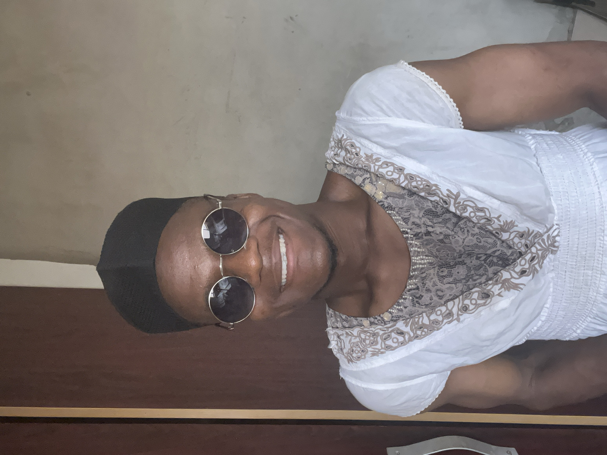 The image, taken in Minna, Nigeria, features a person with dark skin smiling broadly and looking directly at the camera. The photo appears to be a portrait taken upright and then rotated 90 degrees counter-clockwise. The subject is wearing a black head covering, possibly a kufi or skullcap, and round sunglasses with dark lenses and metal frames.
They are dressed in a white short-sleeved top that features intricate decorative patterns in brown and white around the neckline and shoulders, as well as smocking or ruching details on the side. The person's bright, wide smile reveals white teeth, conveying a sense of happiness and friendliness.
The background consists of a plain, light-colored wall on the left side of the rotated image and a dark brown surface, likely a wooden table or piece of furniture, on the right. A thin golden trim line separates the wall from the brown surface. A cropped hint shows a silver-colored handle on the dark brown surface, suggesting it might be a drawer or cabinet. The lighting is soft and even, indicating an indoor setting. No visible text is present in the image.