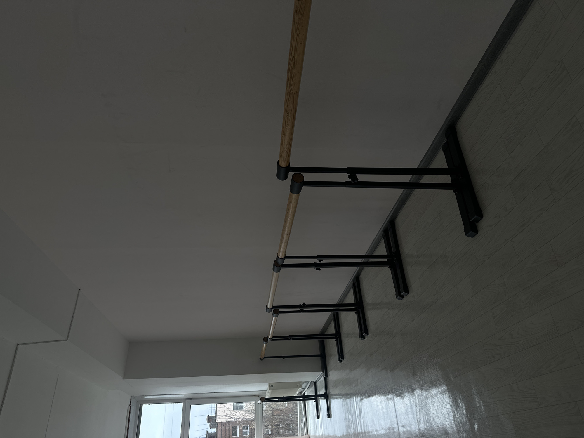 This image depicts a room, potentially a dance studio, with a ballet barre mounted along one wall. The barre has a wooden rail supported by black metal brackets. The wall is light-colored and appears to be made of panels. There is a window on the left side of the image, through which buildings can be seen. The room is located in the city of Votkinsk, Russia.