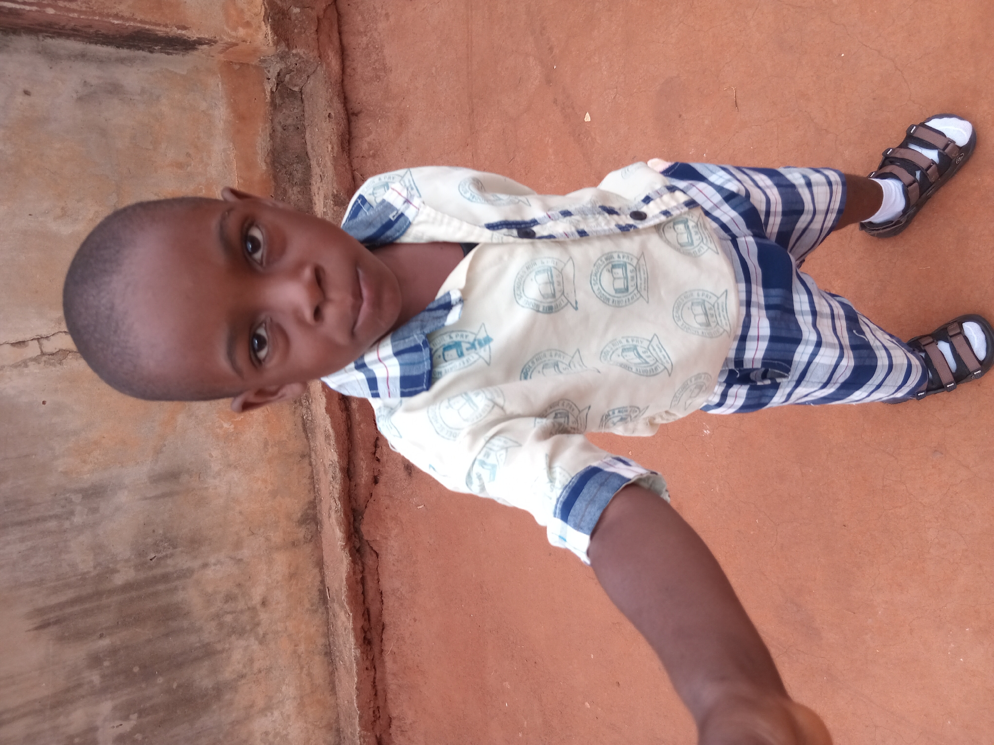 A young boy is the subject of the photograph. He has short, dark hair and dark eyes. He is wearing a light-colored button-down shirt with blue trim and logos, blue and white plaid shorts, and brown sandals with white socks. The boy is standing on a reddish-brown surface, likely dirt or a painted area. Adjacent to him is a textured gray surface, possibly a wall or walkway. The image seems to be taken from a higher angle, looking down at the boy. The location is stated to be in Shagamu, Nigeria. No other individuals, objects, or interactions are explicitly visible in the image.