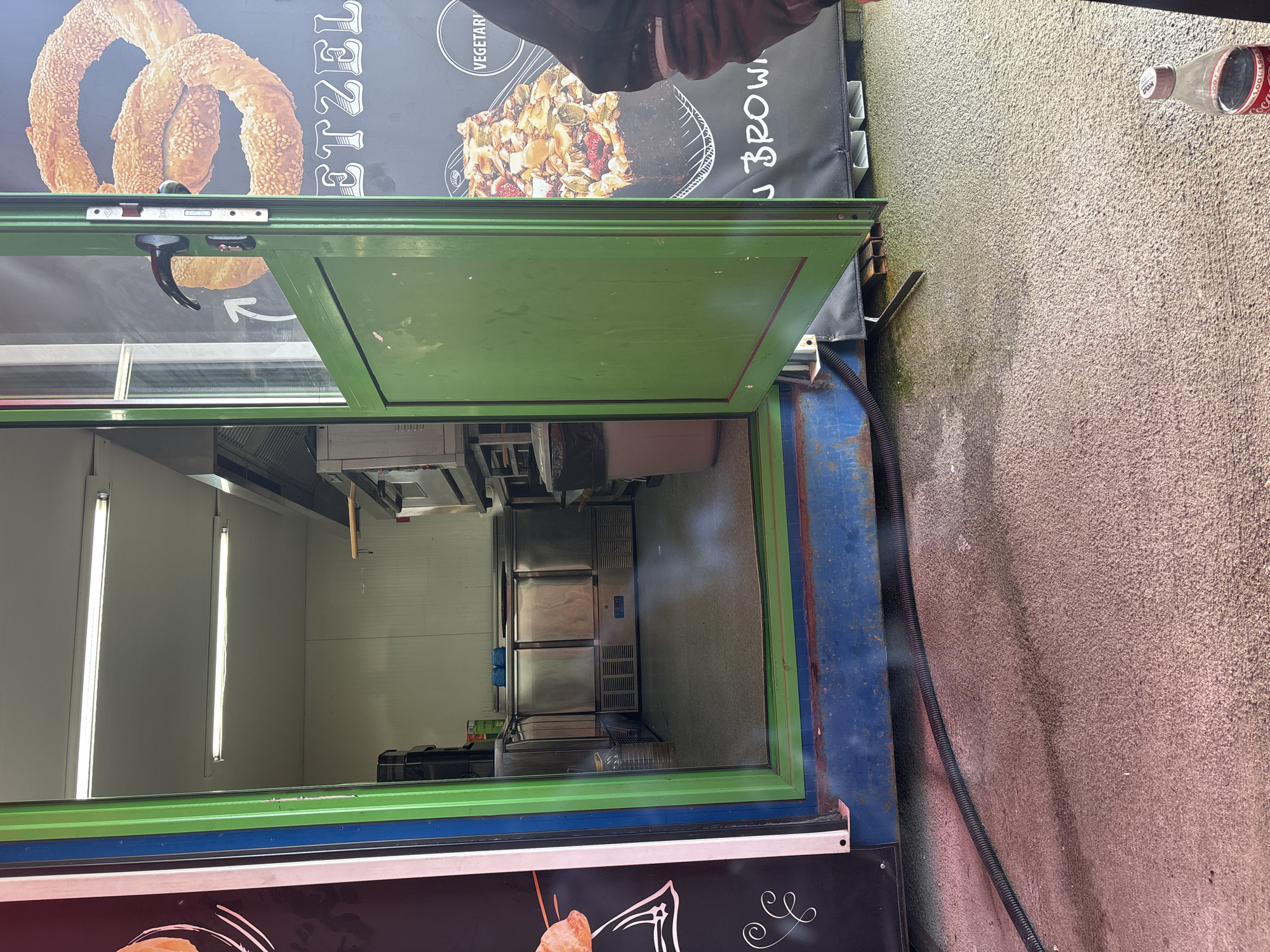 The image shows the open door of a food stall or mobile kitchen, revealing the interior. The stall is located outdoors, with a rough concrete surface visible to the right. The door is painted green, and the exterior of the stall is adorned with promotional graphics featuring food items like pretzels and brownies. Inside the stall, stainless steel equipment such as refrigerators and preparation areas are visible. Two fluorescent lights are affixed to the white interior walls, indicating a hygienic workspace. A black flexible conduit is present along the exterior wall, likely providing electrical power. The image provides no information about the stall being located in Burgas, Bulgaria.