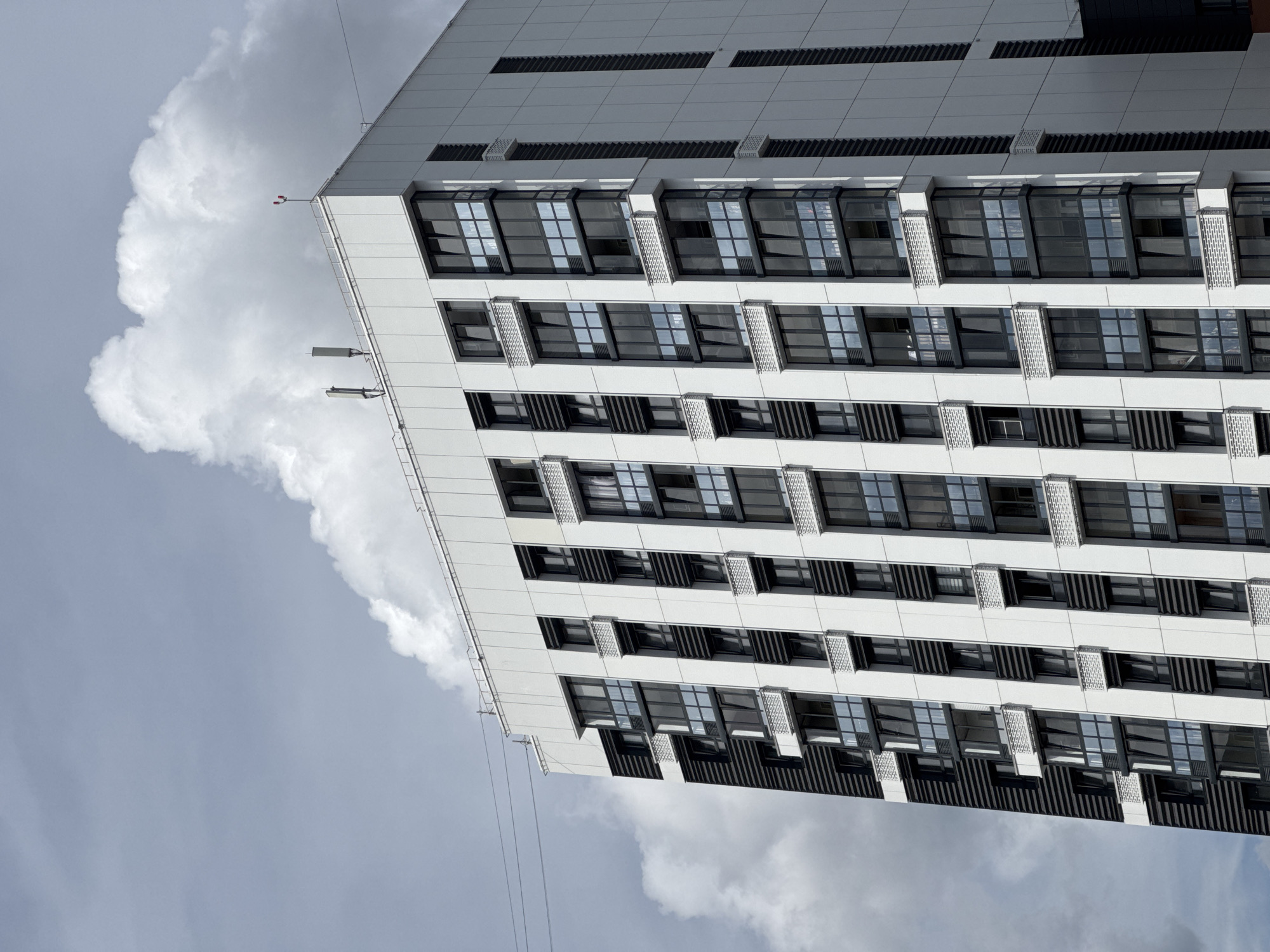The image captures the upper section of a modern, multi-story apartment building with a predominantly white facade accented by black vertical elements and dark-framed windows. The building features repeating patterns of glassed-in balconies or window sections. The roofline is visible, with what appear to be two antennas and a small red and white beacon. The sky is cloudy, with a large, billowy white cloud dominating the upper right portion of the frame, partially obscuring a bright blue sky. Subtle power lines are visible in the upper left quadrant, stretching across the frame. There is no discernible human activity. The architectural style suggests a contemporary urban residential development. The image is framed from a low angle looking upwards.