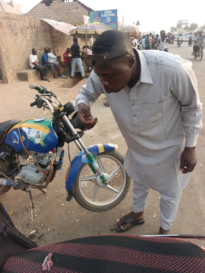 An outdoor scene in Alizaga, Nigeria, depicts a man in a light-colored traditional tunic and trousers, with sunglasses perched on his head, leaning over a blue and yellow decorated motorcycle parked on its kickstand. He is observing a small, dark object held in his left hand. His right hand is at his side, and he wears dark sandals. The motorcycle features a five-spoke silver front wheel and various decals, including a visible Nigerian flag motif on its tank. The immediate ground is unpaved and dusty, transitioning to a paved road further into the background. To the left, several men are seated on a low concrete wall. The background reveals a bustling street environment with multiple additional motorcycles, pedestrians, and market stalls under umbrellas, one displaying "SEEING IS BELIEVING GOMBE 2027." Overhead electrical lines and a utility pole are also visible. The scene is illuminated by daylight.