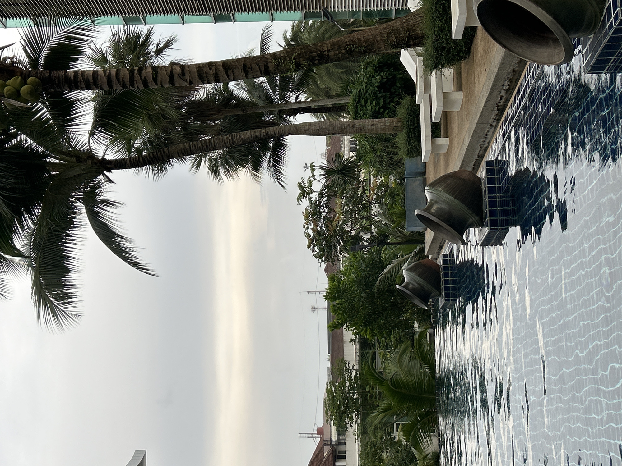 A swimming pool with dark blue tile lining and a rippling surface fills the foreground of the image. Two large, dark, overturned urn-shaped decorative objects are placed on tiled pedestals at the edge of the pool. To the right of the pool, a concrete walkway is lined with white lounge chairs and low white benches. Beyond the walkway, lush green foliage and palm trees create a tropical backdrop. In the distance, the roofs of buildings and telecommunication towers are visible under an overcast sky, suggesting an urban or suburban setting in Kathu, Thailand. A modern building with a green, ribbed facade is visible on the far right. The scene appears to be an outdoor common area, possibly at a hotel or residential complex.