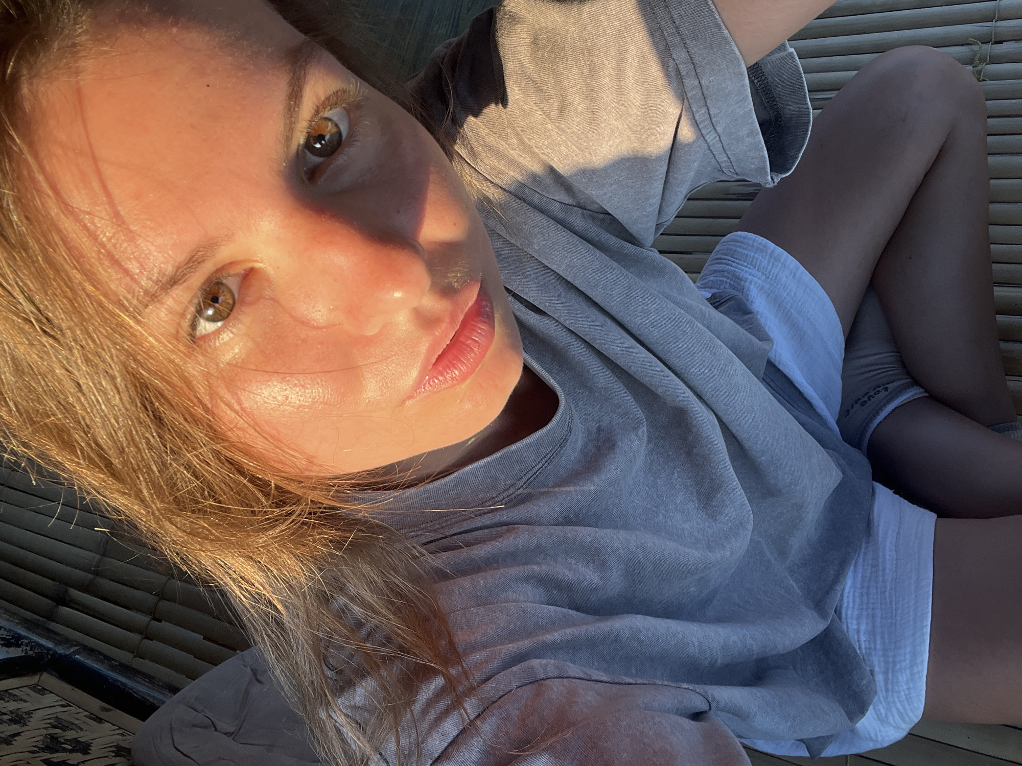 In Kertasari, Indonesia, a young woman with auburn hair takes a selfie. She is seated on a woven bamboo surface, her body angled towards the camera. Sunlight casts a warm glow across her face, highlighting her brown eyes and parted lips. She wears a grey t-shirt and white shorts. To her left, a grey pillow rests against a dark, textured surface, possibly a wall or partition. The lighting suggests it is either early morning or late afternoon.