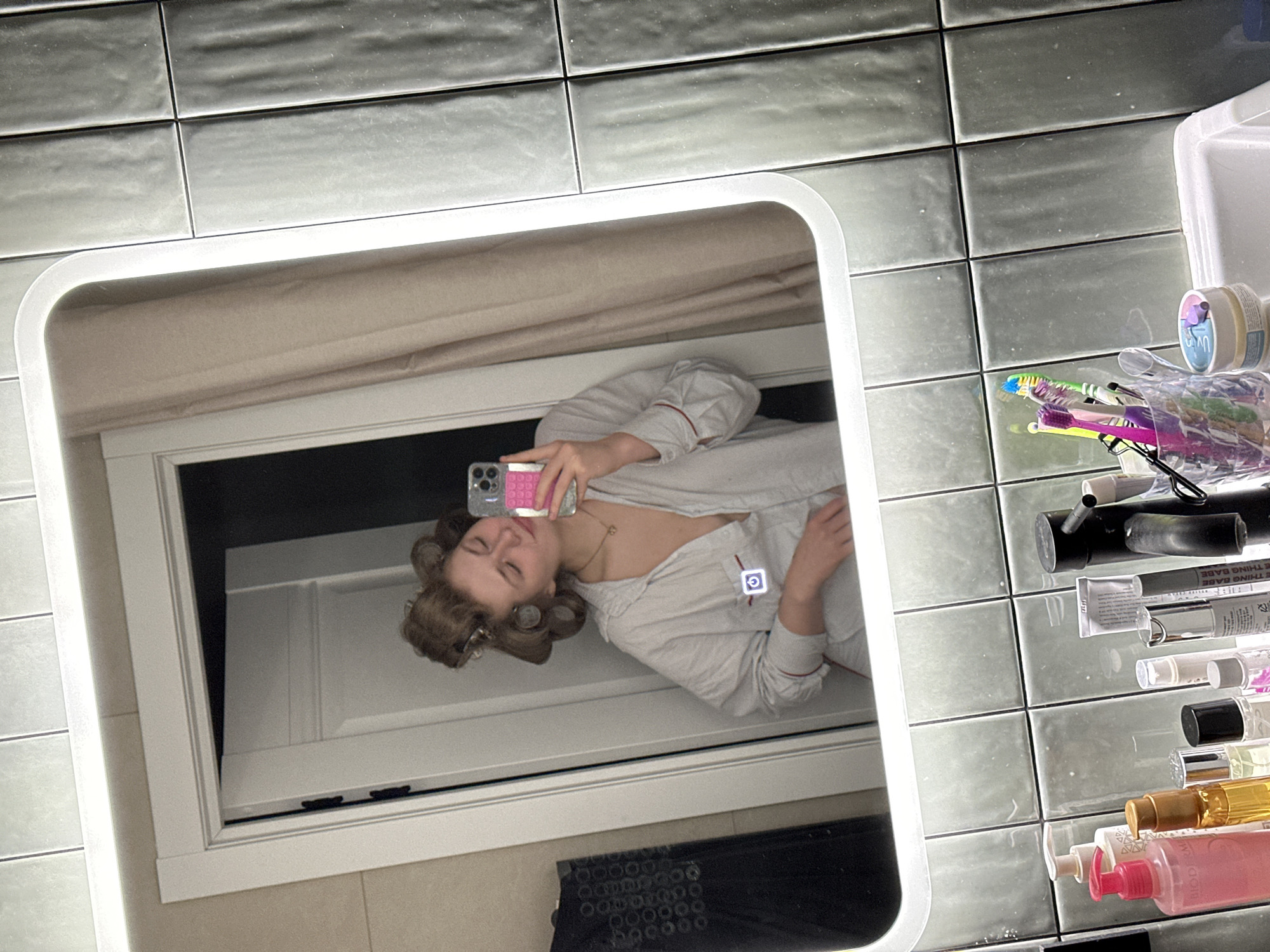 The image shows a bathroom scene, likely taken by a mirror selfie. A woman with hair curlers is seen in the mirror, holding a phone with a pink case. She is wearing a light-colored top. The mirror reflects a door behind her. The surrounding wall is tiled in grey. To the right of the mirror, various bathroom items are visible, including toothbrushes, makeup, and skincare products. There is a container and a shelf with several bottles and tubes. The location is likely a bathroom, but the city of Kazan, Russia, cannot be determined from the provided image.