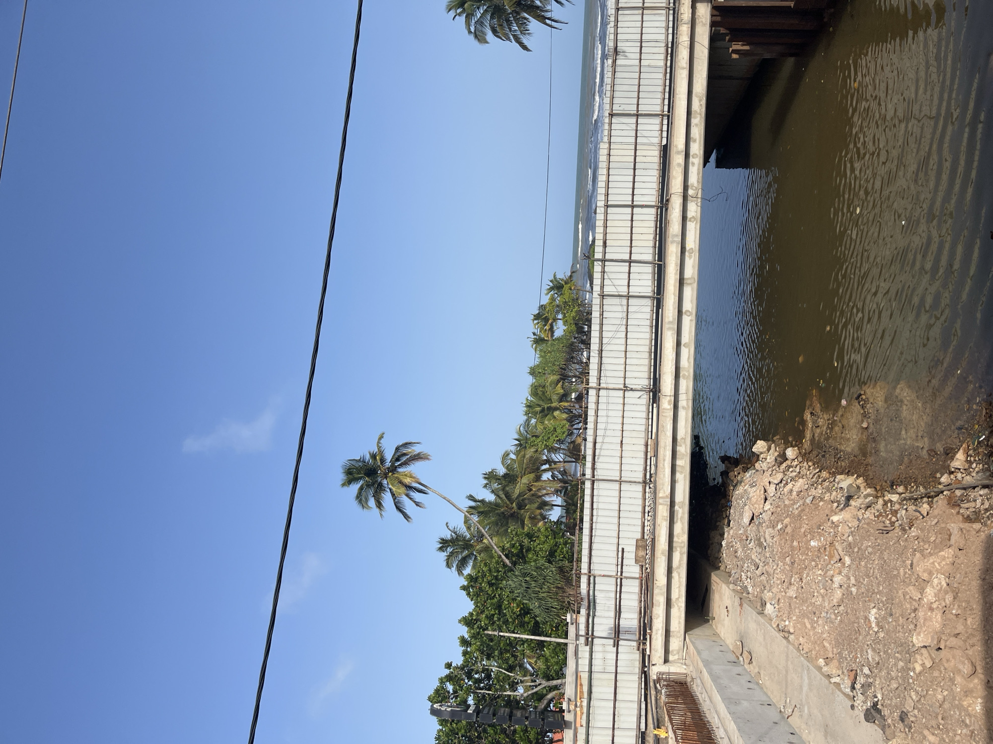An eye-level, medium outdoor shot captures a scene of ongoing construction on a bridge over a small body of water in Maggona, Sri Lanka. The sky is a clear, bright blue, with a few wispy clouds and two black power lines stretching across the upper portion of the frame.
In the foreground, the body of water appears murky and brown, with ripples on its surface. The water's edge on the left is lined with rough, rocky terrain and some construction debris. A concrete pathway runs alongside the water and the construction site.
The bridge itself is a light-colored concrete structure, supported by concrete piers. A temporary metal barrier, likely for construction, runs along the top edge of the bridge. Underneath the bridge, the water flows into a darker, more shadowed channel.
Behind the bridge, a dense line of lush green trees and palm trees creates a tropical backdrop. One particularly striking palm tree leans dramatically to the left, its fronds silhouetted against the sky. In the distance, the ocean can be seen, with gentle waves rolling towards the shore. The overall impression is of a coastal area undergoing development.