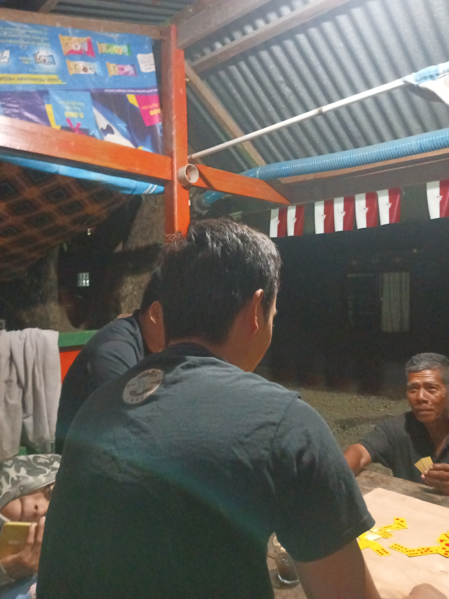 In the city of Tumampua, Indonesia, a dimly lit, open-air gathering takes place under a corrugated metal roof supported by orange wooden beams. The scene depicts at least three individuals engaged in a game of dominoes. In the foreground, the back of a man wearing a black t-shirt is visible. To his right, an older man in a black t-shirt is seated at a wooden table, holding dominoes. In the background, another individual is partially visible, also in a dark shirt. A younger person wearing a camouflage hat is on the far left, holding a smartphone. The table is set with yellow domino tiles arranged in a pattern, indicating a game in progress. The environment appears to be rustic, with visible wiring and a patterned tarp or banner on the left wall. Red and white striped banners hang from the ceiling. The lighting is dim and artificial, creating shadows and highlighting the activity.