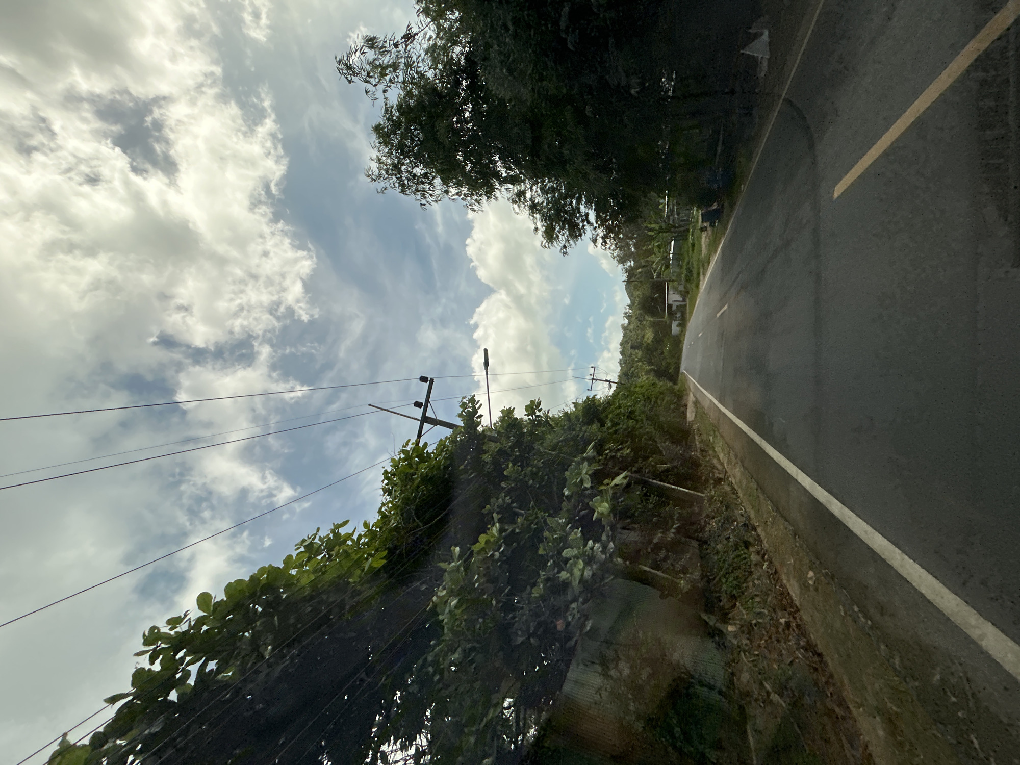 The image shows a view from a vehicle traveling on a two-lane asphalt road in a rural area, likely in the city of Ratsada, Thailand. The road is lined with lush green trees and vegetation on both sides. Overhead, power lines stretch across the frame, supported by utility poles. The sky is partly cloudy with scattered white clouds against a blue backdrop. In the distance, small buildings and some houses are visible, suggesting a sparsely populated area. The road has a white line marking the edge on the left and a yellow line marking the center. The overall scene depicts a typical rural landscape with infrastructure elements like power lines.