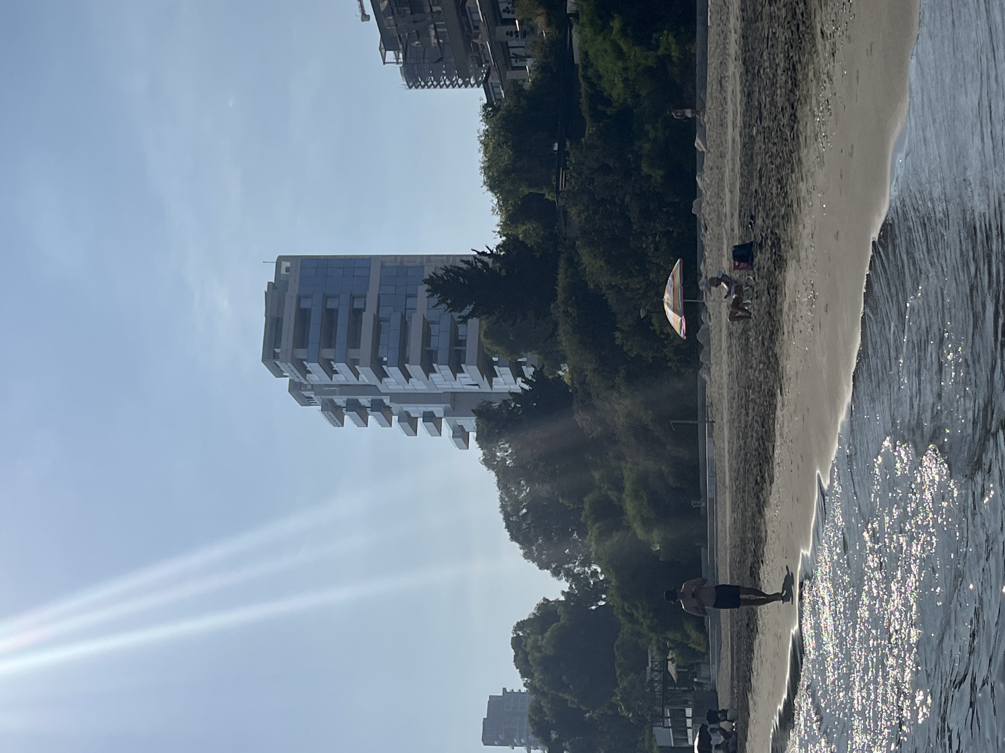 A sun-drenched beach scene in Agios Tychona Community, Cyprus, showcases a tranquil day with a few individuals enjoying the shoreline. In the foreground, gentle waves lap against the sandy beach, reflecting the bright sunlight. A man in dark shorts and a hat is walking along the wet sand, appearing to be looking at his phone. Further down the beach, an umbrella provides shade for a person sitting in a chair, also engaged with a mobile device. In the background, lush green trees and vegetation form a backdrop to modern apartment buildings with balconies. The clear blue sky is visible, with distinct rays of sunlight streaking across the upper left portion of the image. The overall atmosphere is one of peaceful relaxation by the sea.