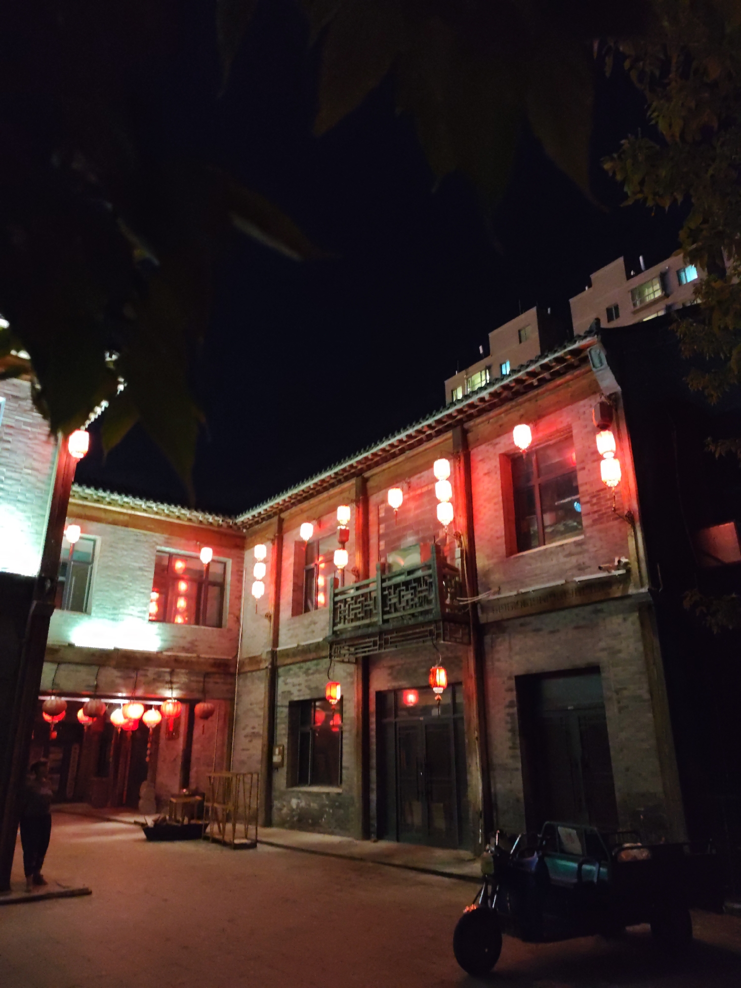 A night-time scene in Lindian, China, features a traditional Chinese courtyard building illuminated by numerous red lanterns. The building, constructed of brick, has multiple windows and a balcony adorned with intricate carvings. In the foreground, a person stands near the entrance to the courtyard, and a three-wheeled vehicle is parked on the ground. In the background, modern apartment buildings rise above the traditional architecture. The overall atmosphere is one of vibrant cultural celebration set against a backdrop of urban development.