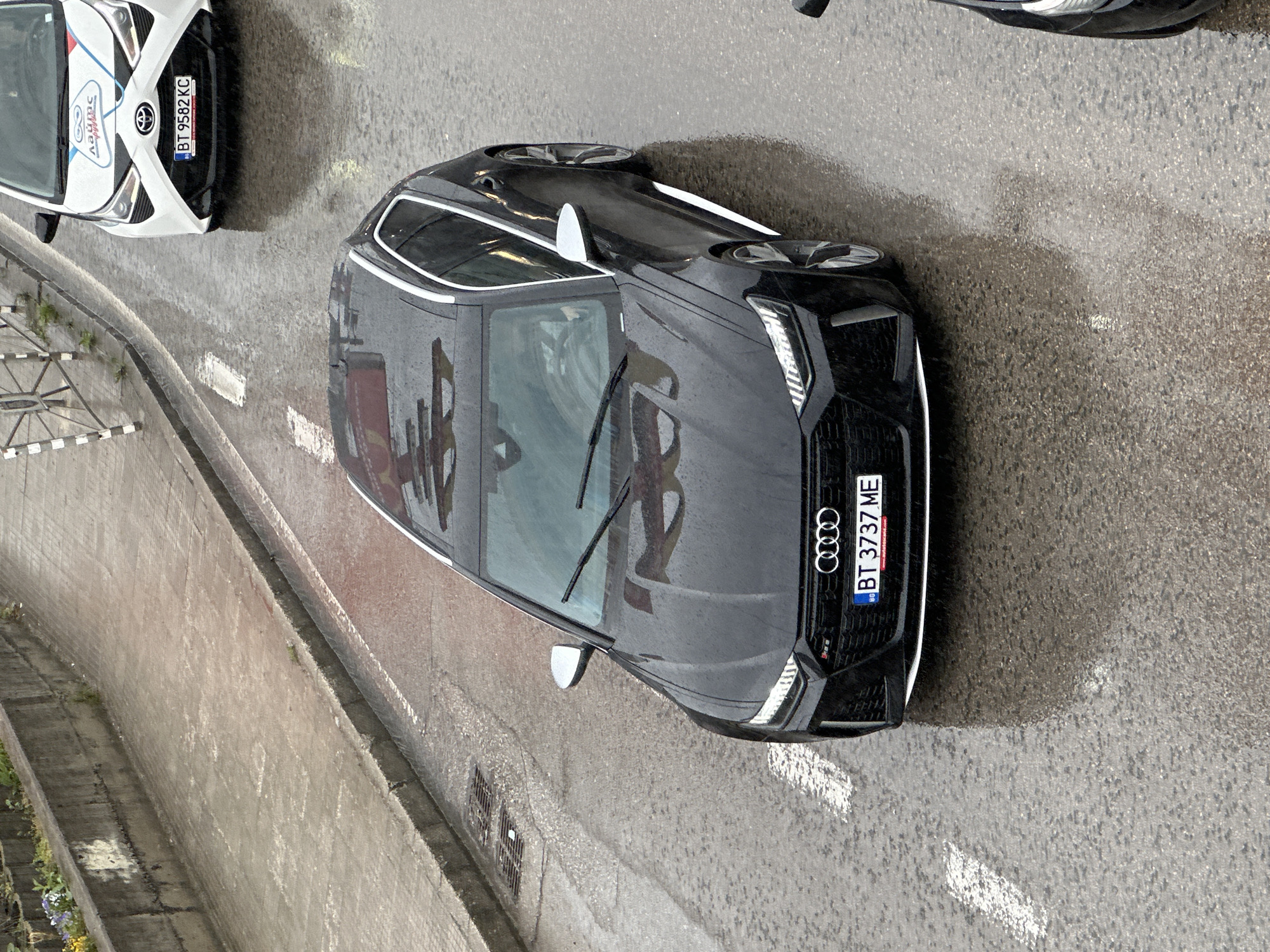 The image shows a black Audi RS6 parked on a wet road in Veliko Tarnovo, Bulgaria. The license plate reads "BT 3737 ME." The car is oriented towards the right of the image and reflects the surrounding buildings on its hood and windshield. Adjacent to the Audi is a white Toyota Aygo with the license plate "BT 9582 KC." Both cars are parked alongside a concrete barrier, with white road markings visible on the asphalt. The overall scene suggests a rainy day in an urban environment.