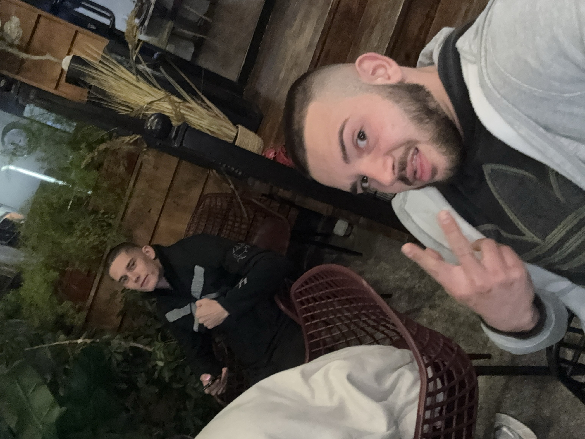 Two men are pictured in an outdoor setting. One man is holding up a peace sign, wearing a gray hoodie with a dark shirt. His head is shaved and he has a beard. The other man is wearing a dark jacket with lighter-colored stripes and looking directly at the camera. Both men are sitting on brown, woven-style chairs. The background includes greenery, a light fixture with bundled straw, and wooden structures. The location is likely Tsarevo, Bulgaria, based on the question context.