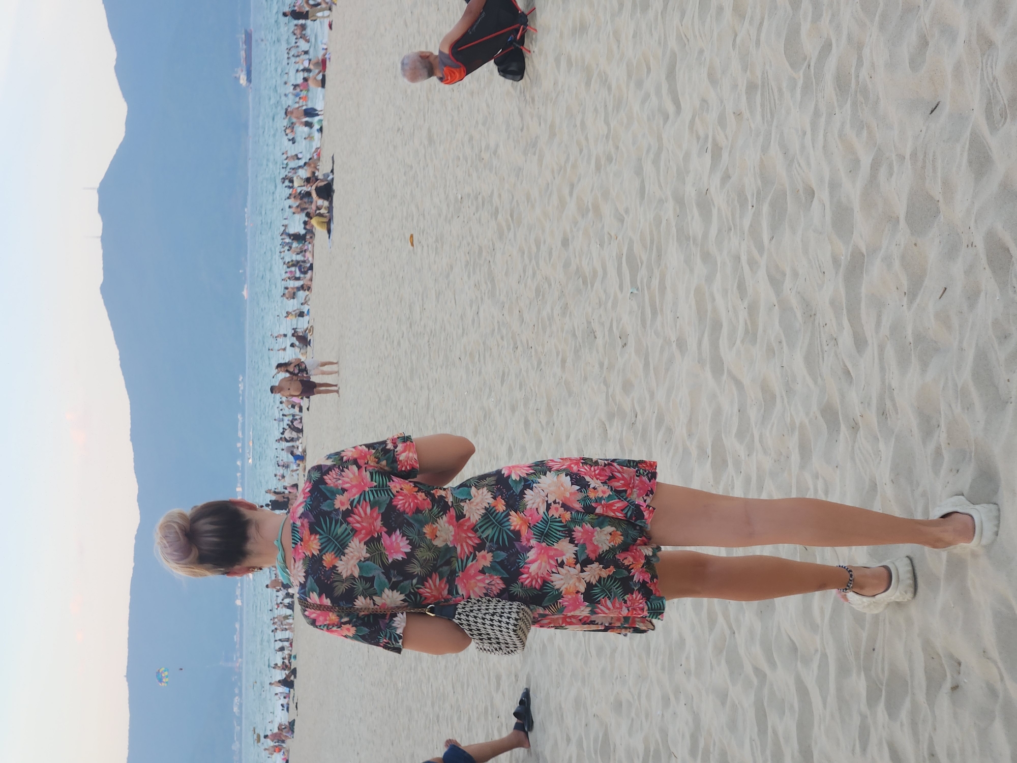 The image depicts a busy beach scene in Da Nang, Vietnam. The primary subject is a woman from behind, wearing a floral-patterned dress and white shoes. She is standing on a sandy beach with many other people visible in the background, suggesting a popular tourist destination.
The scene is set by the ocean, with mountains forming a backdrop on the left side of the frame. The water is blue and appears calm, with a boat on the horizon and a parasailer in the distance. The beach itself is made of fine, light-colored sand, with footprints and ripples indicating recent activity.
The time of day appears to be late afternoon or early evening, judging by the soft light and the colors of the sky, which is a pale blue. The weather seems pleasant, as many people are out enjoying the beach, some in the water, others walking or relaxing on the sand. The atmosphere seems lively and relaxed, with a sense of vacation and leisure.
Several other people are visible scattered across the beach, some partially obscured. One person in the upper part of the image is wearing an orange and black shirt. The overall impression is a vibrant, sun-drenched beach teeming with life. No visible text is present in the image.