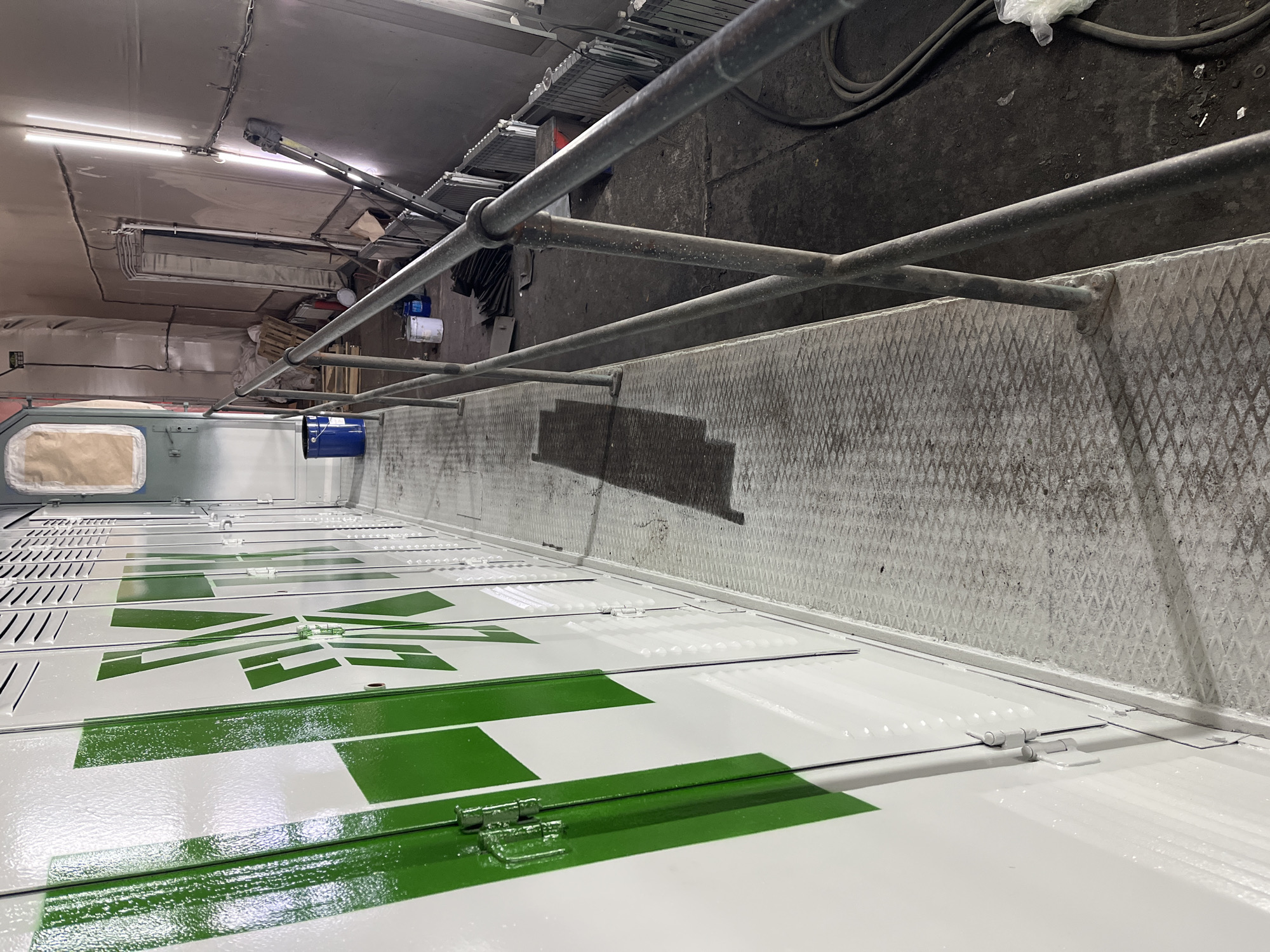 The image shows an interior shot of a vehicle, likely a truck or trailer, in an industrial setting in or around Chaikovsky, Russia. The interior is dominated by white panels with green geometric shapes and lines. Some panels have hinges. A metal railing system runs along one side. The floor is concrete or metal with a diamond plate pattern. Overhead, there are industrial fixtures, including lighting and possibly some scaffolding or storage racks. The area appears to be a workshop or storage space.