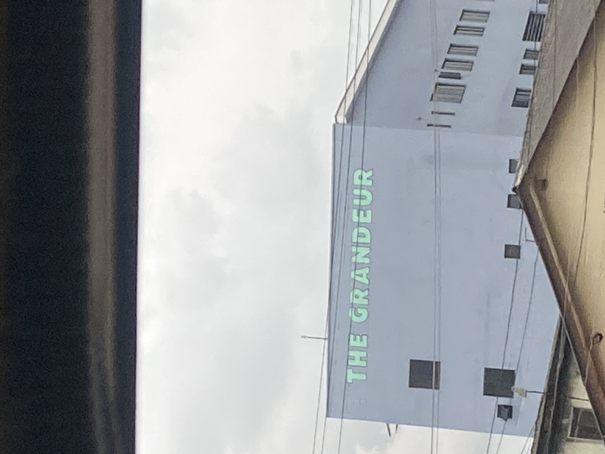 The image shows the upper facade of a light blue building with "THE GRANDEUR" written in large, light green, sans-serif font across its upper section. The building is located in Ikeja, Nigeria. Several windows are visible on the facade, some appearing dark, and others with blinds. Power lines run horizontally across the building. In the foreground, the sloped roof of a lower, older building is visible, with its facade partially obscured. The sky above is overcast.