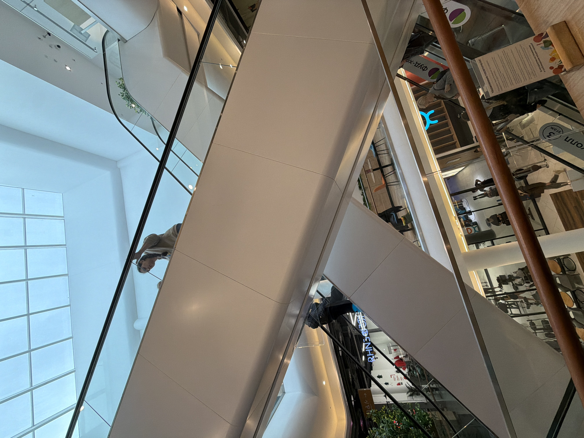 The image presents an interior view of a shopping center, possibly in Moscow, Russia, given the Cyrillic text in the reflections. The scene is dominated by a modern architectural design featuring white structural beams, glass panels, and escalators. The camera is angled upwards, emphasizing the multi-level construction of the building. Reflections in the glass reveal interior details, including shops and people. A woman is visible in one reflection, riding an escalator. There are plants visible on one of the floors of the shopping center.