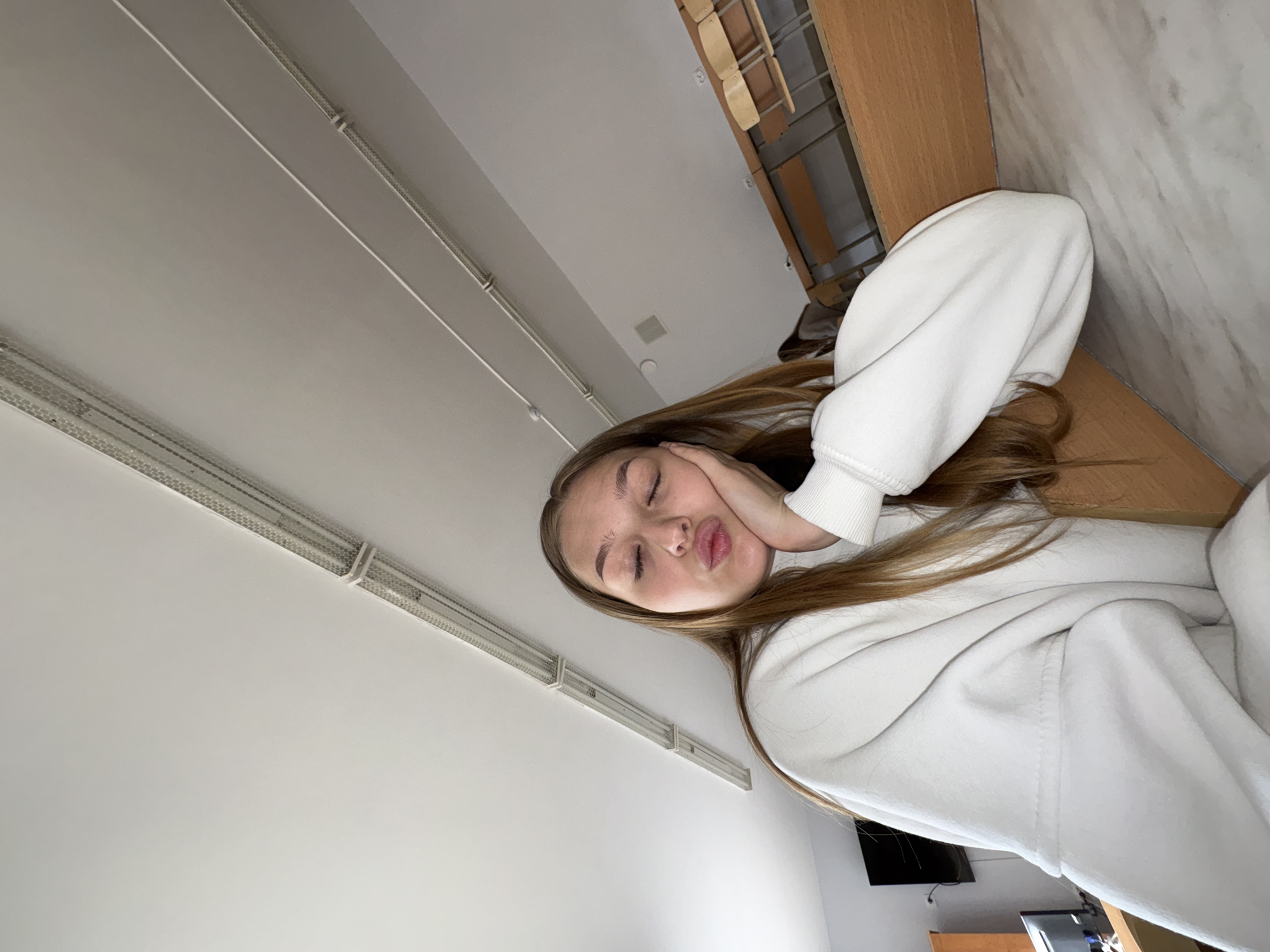 In a classroom or lecture hall in Chaikovsky, Russia, a young woman with long, light brown hair is pictured in a close-up, selfie-style shot. She is wearing a white hooded sweatshirt. Her eyes are closed, and her lips are puckered as if she is about to kiss or has just kissed. Her right hand is resting on her cheek, supporting her head. The background includes fluorescent lighting fixtures on the ceiling, wooden desks with chairs, and a wall-mounted television screen in the distance. The overall impression is one of a casual moment captured during a study session or a break.