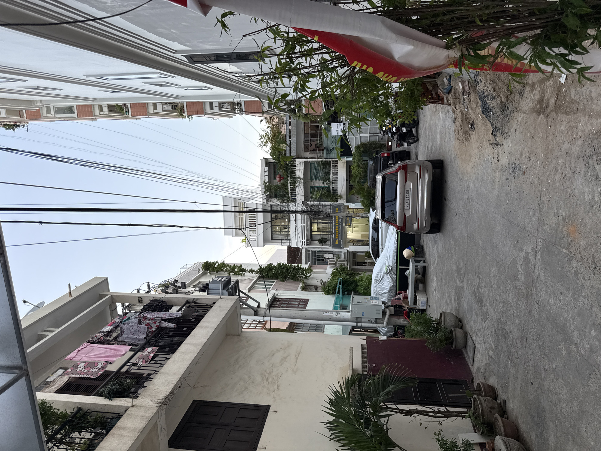 The image shows a narrow street or alleyway in Nha Trang, Vietnam, with multi-story buildings lining both sides. The street is paved with concrete and appears to be residential or mixed-use.
On the left side of the street, a light-colored building features a balcony with laundry hanging out to dry, including pink and patterned clothing. A dark-colored window is visible on the lower floor. Several potted plants are placed along the base of this building.
On the right side of the street, taller buildings are visible, some with balconies and greenery. A white building with red brick accents is prominent. Several utility wires crisscross overhead.
In the middle of the street, two cars are parked. The car in the foreground is a silver SUV with the license plate "79A-614.08". Another car, partially covered with a silver tarp, is parked behind it. A motorcycle is parked further down the alley.
A large red and white fabric banner or flag is draped on the right side of the frame, partially obscuring a dense cluster of bamboo-like plants. The overall impression is of a dense urban environment with signs of everyday life.