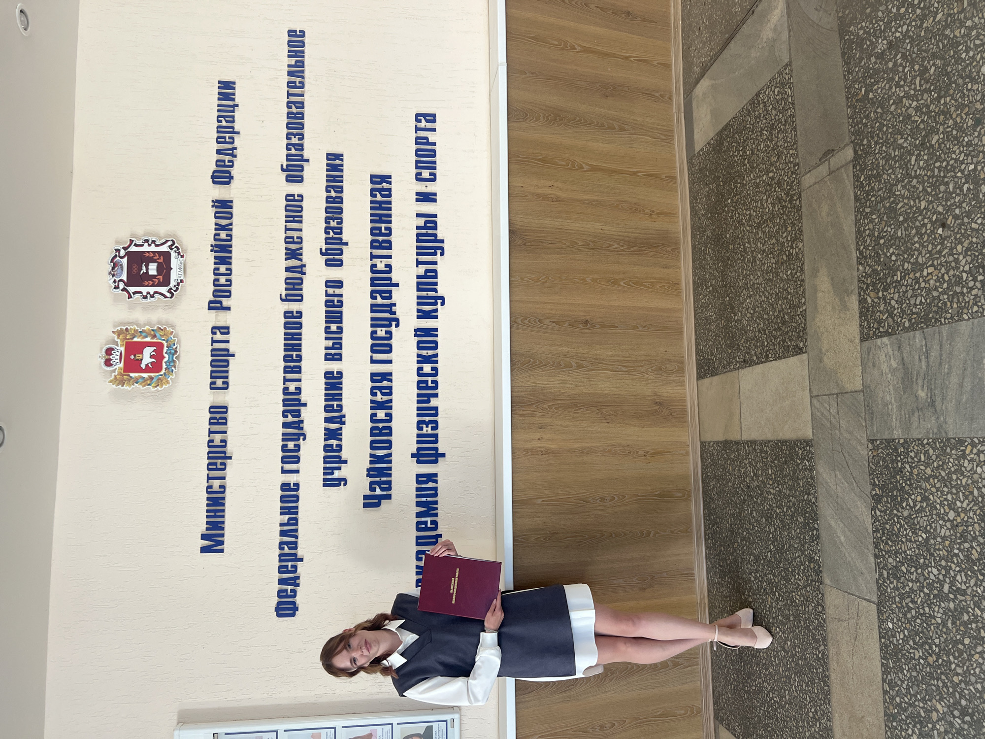 A young woman stands in front of a wall with blue lettering and emblems. She is wearing a white shirt, a grey vest, and a white skirt, along with light pink heels. She is holding a dark red folder in her left hand. The wall behind her has the words "МИНИСТЕРСТВО СПОРТА РОССИЙСКОЙ ФЕДЕРАЦИИ" (Ministry of Sport of the Russian Federation) and below it "ФЕДЕРАЛЬНОЕ ГОСУДАРСТВЕННОЕ БЮДЖЕТНОЕ ОБРАЗОВАТЕЛЬНОЕ УЧРЕЖДЕНИЕ ВЫСШЕГО ОБРАЗОВАНИЯ" (Federal State Budgetary Educational Institution of Higher Education). Underneath that, it reads "ЧАЙКОВСКАЯ ГОСУДАРСТВЕННАЯ АКАДЕМИЯ ФИЗИЧЕСКОЙ КУЛЬТУРЫ И СПОРТА" (Chaikovsky State Academy of Physical Culture and Sport). The floor is tiled with large, square grey and speckled sections. The wall below the lettering is covered in wood-paneled material.
