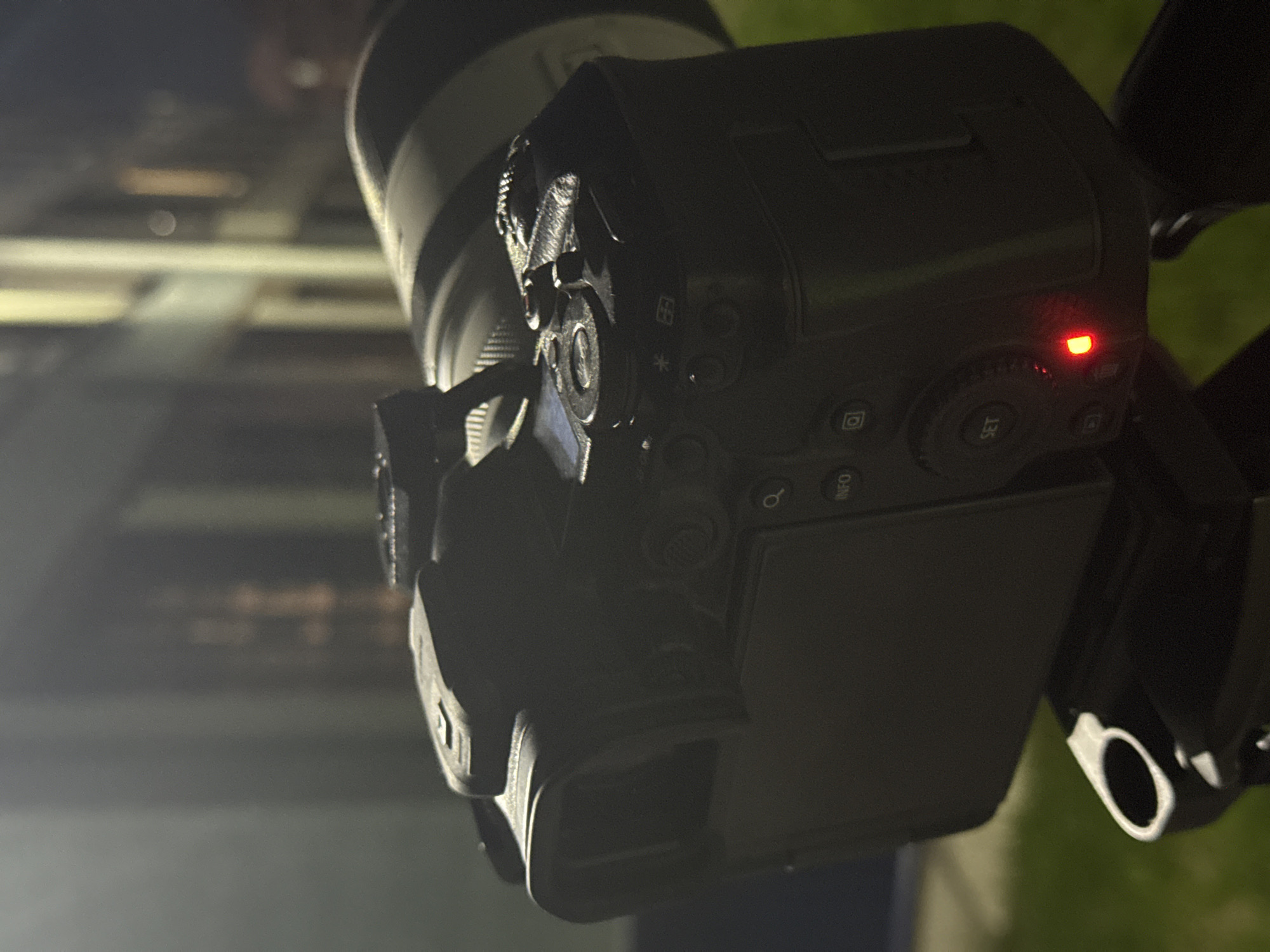 The image shows a digital camera on a tripod. A red light is illuminated on the back panel. The camera is pointed toward a blurred background with horizontal lines suggesting buildings in a city. The location cannot be specifically identified as Perm, Russia without further information.