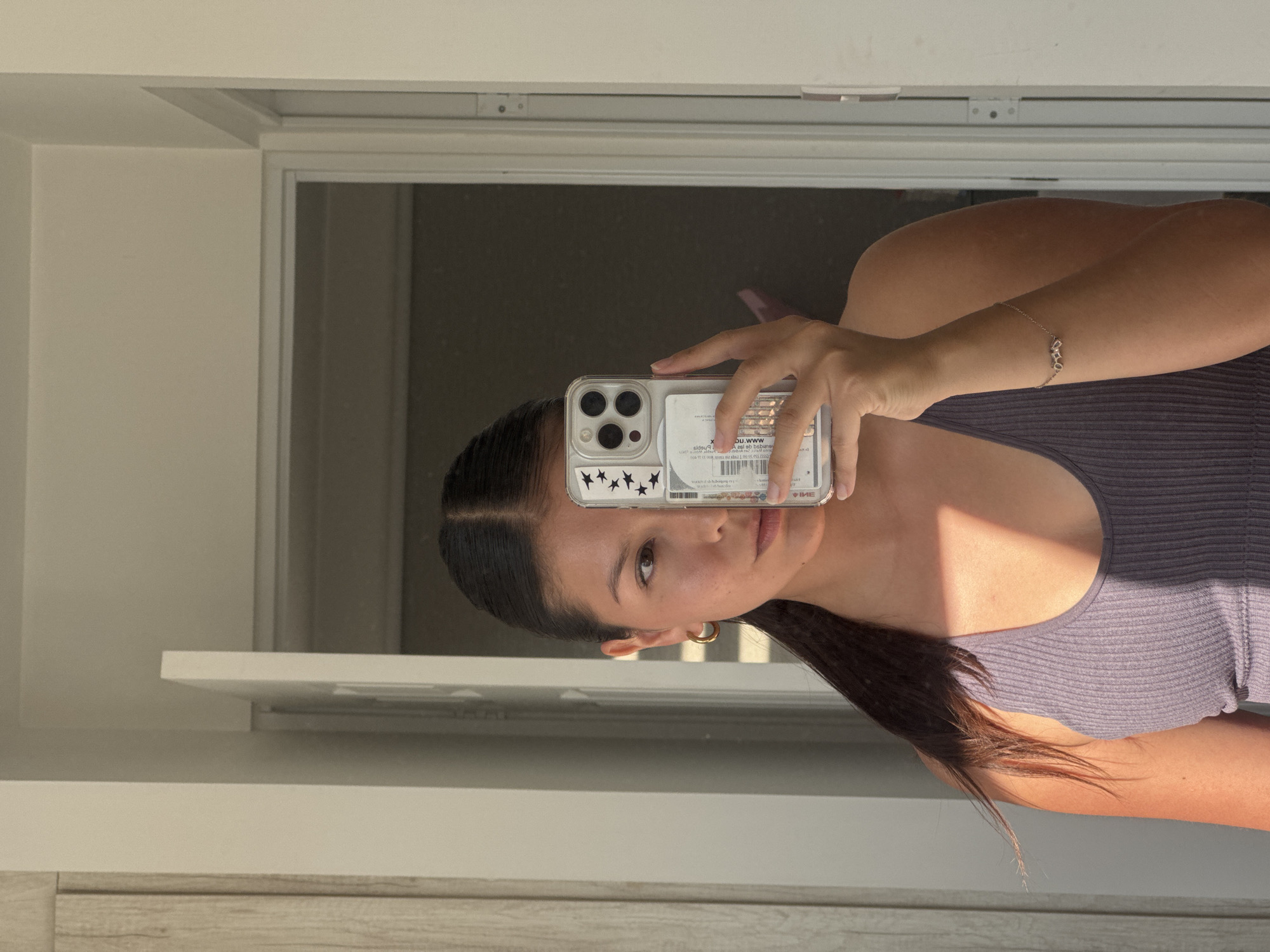 The image is a mirror selfie, rotated 90 degrees clockwise, depicting a young person indoors in San Francisco Ocotlán, Mexico. The person, likely female, has dark hair styled with a middle part and is wearing a light purple ribbed tank top. They hold a clear smartphone with a white bumper, featuring a sticker with black stars on the lower left of the case. Through the transparent back of the phone case, several cards or documents are visible, displaying mirrored and partially legible text, including fragments like "WWW.D.DU" and "IME," along with numbers.
The person's right hand holds the phone, while their left arm, adorned with a delicate bracelet featuring small geometric shapes, is also visible. They look directly at the camera with a neutral expression. The indoor setting features light-colored walls and a doorframe. Strong natural light, consistent with sunshine, casts distinct shadows on the person's right shoulder and arm, indicating it is daytime, possibly morning or late afternoon. A light-colored counter or shelf is visible below the mirror, with a wooden-textured surface beneath it.