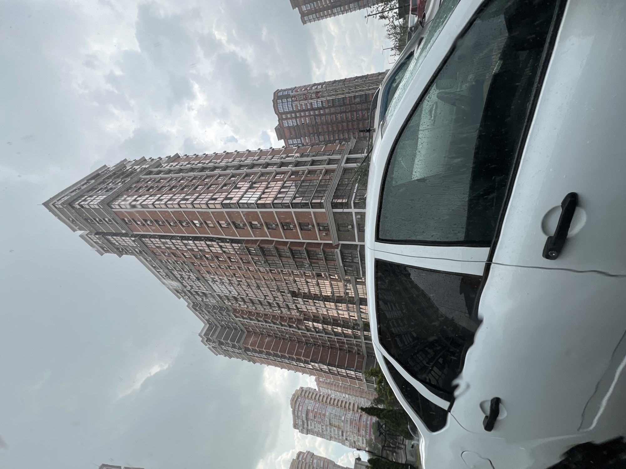The image captures a view of tall, modern apartment buildings in Krasnodar, Russia. The buildings are characterized by their brick facades and numerous windows, some of which appear to be tinted. In the foreground, a white car is partially visible, with water droplets on its windows, suggesting it is raining or has recently rained. The sky is overcast with clouds, indicating a cloudy or potentially stormy day. The perspective is from inside or next to the car, looking upwards towards the buildings, creating a sense of scale and height. The overall scene conveys a residential urban environment during inclement weather. There is no visible text in the image.