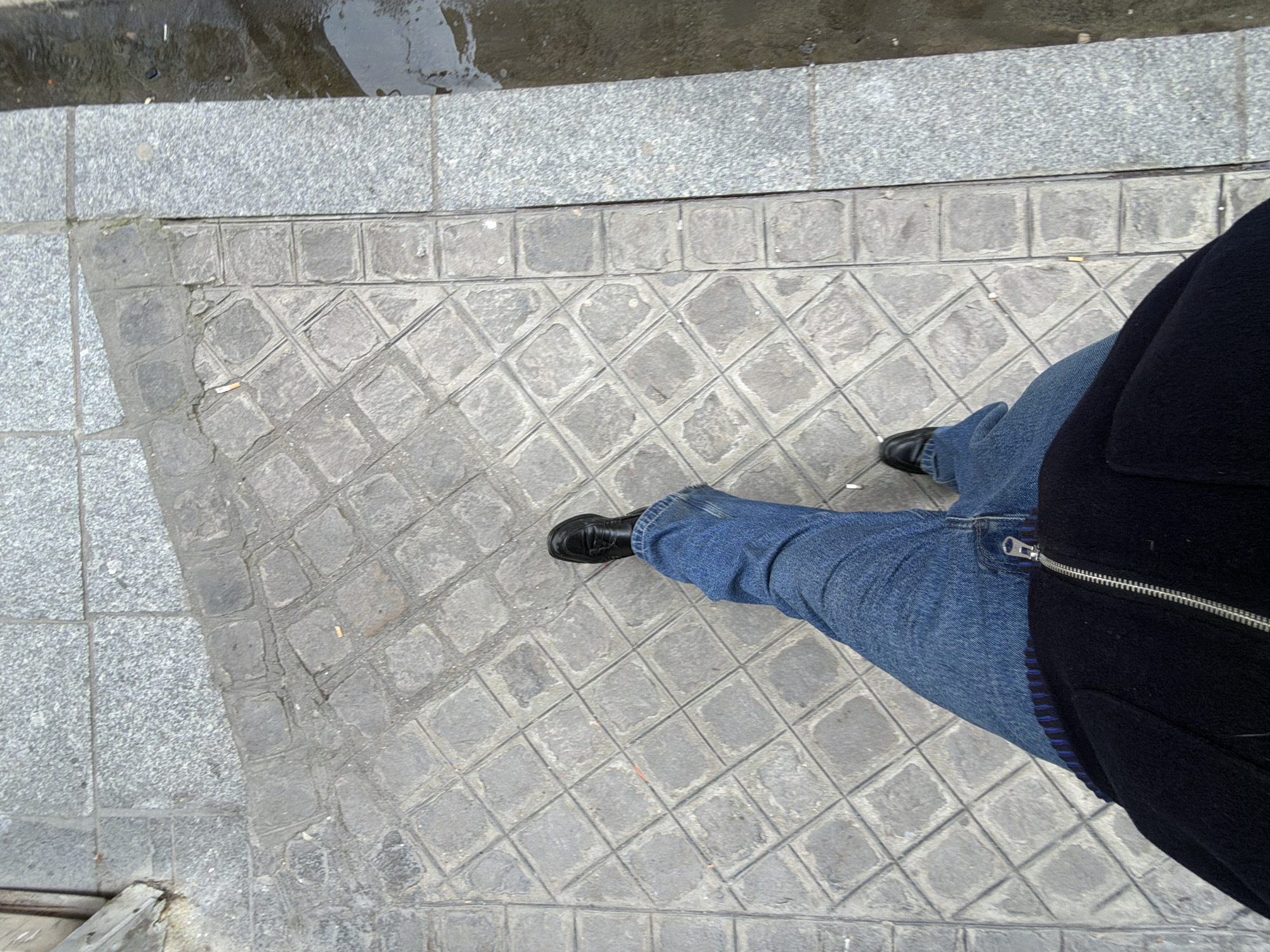 The image shows a person standing on a cobblestone sidewalk in Paris, France. The sidewalk is made of small, square cobblestones arranged in a diamond pattern, bordered by a row of square, but slightly larger stones. The pavement is mostly grey with some darker patches, possibly due to moisture. The person in the image is wearing dark blue jeans and black shoes. A black jacket obscures part of the view. The top left corner of the image reveals a section of grey pavement with a wet area, suggesting the weather may be damp. The photo is taken from a top-down perspective, directly above the person's feet.