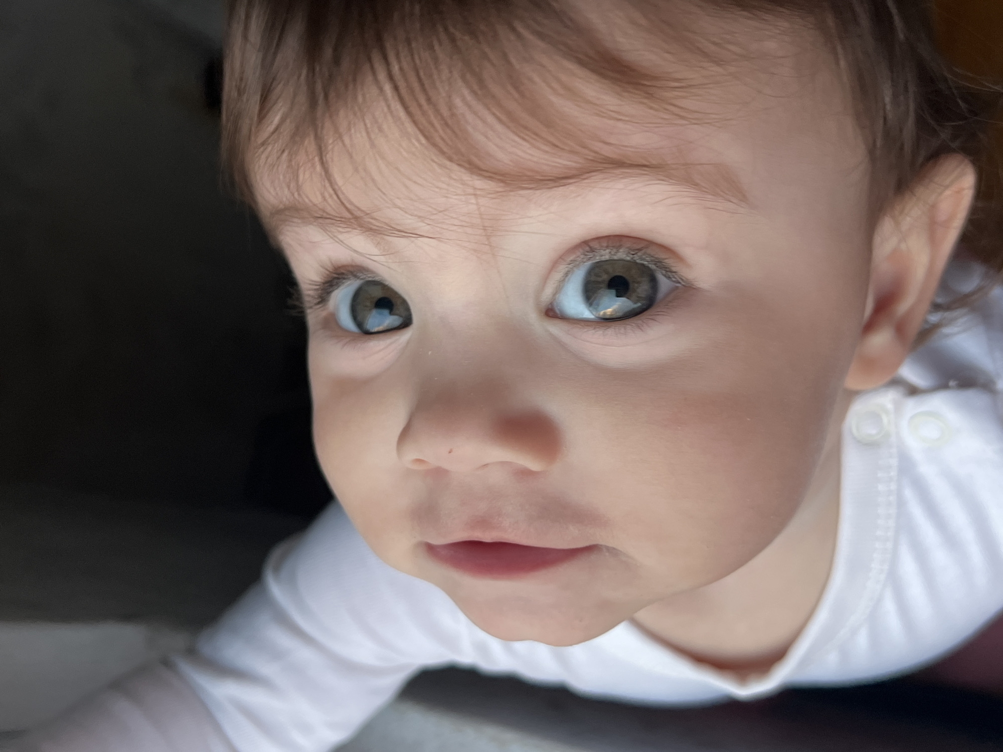 A high-angle, close-up image captures the face of a young child, likely an infant or toddler, looking directly upwards. The child exhibits fair skin and light brown hair, visible along the top and right side of the head. The eyes are prominent, featuring large, light green-hazel irises with distinct reflections in both pupils. The nose is small and upturned, and the mouth is slightly parted, revealing pink lips. The child is wearing a white long-sleeved garment, possibly a onesie, with two visible white snap buttons on the left shoulder area. The background is predominantly dark and out of focus, with a lighter, indistinct area on the far left, suggesting an indoor environment. There are no visual cues within the image to confirm the specified location in the city of Elena, Bulgaria.