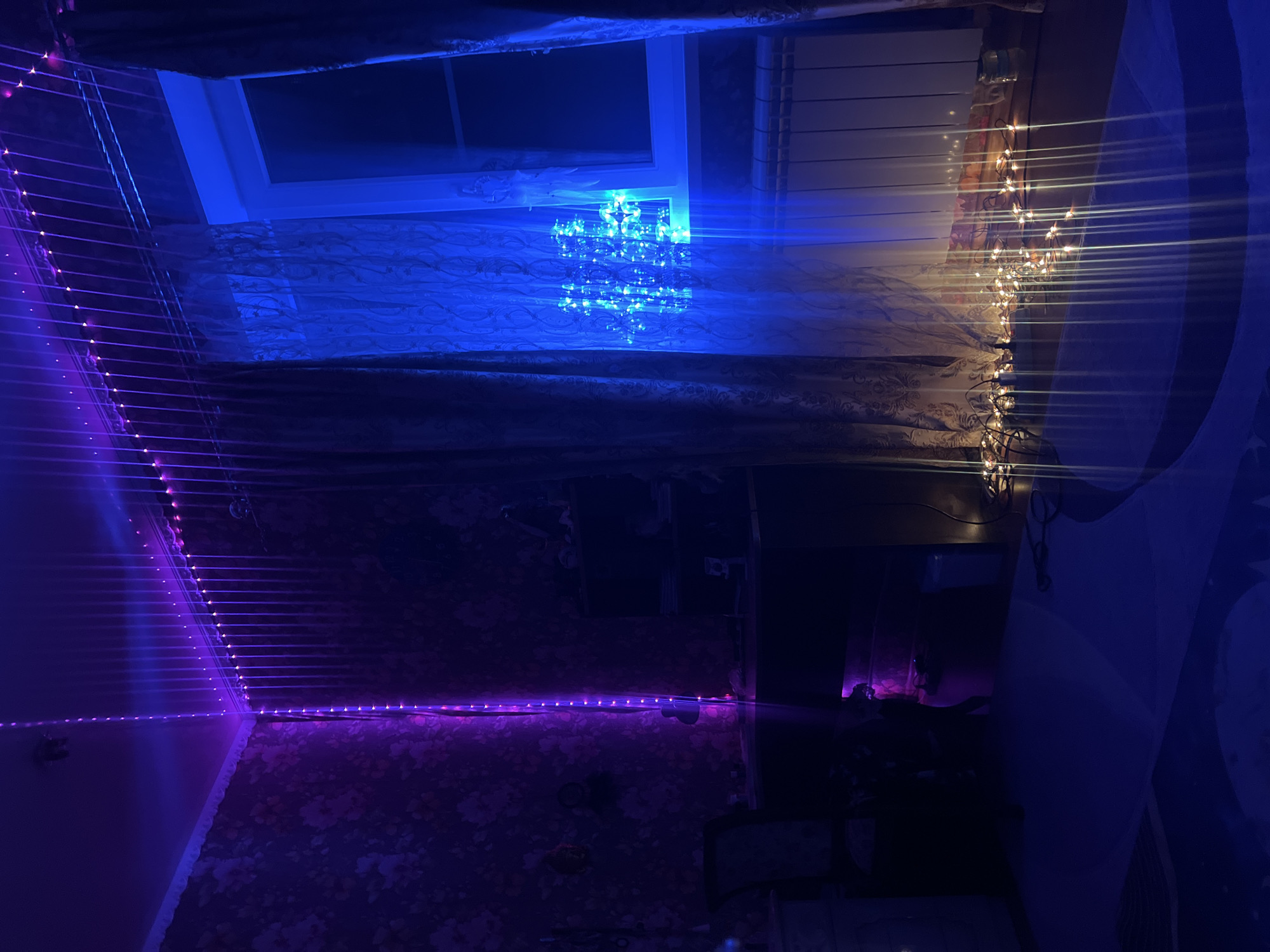 The media file depicts a bedroom interior, illuminated by an assortment of colored LED strip lights and string lights. The dominant colors are deep blue and vibrant purple, creating a moody and atmospheric setting. The camera angle is tilted, giving a sense of being inside the room looking upwards towards a window.
The window is partially obscured by curtains, one set appearing to be sheer and patterned, and another set is darker, likely blackout curtains. Through the window, the night sky is visible, though indistinct. Above the window, blinds are partially drawn. A cluster of bright blue LED lights is positioned on the windowsill, casting a strong glow. To the right of the window, warm-toned string lights are draped, their light creating a starburst effect due to lens flare.
The walls are decorated with a dark, floral-patterned wallpaper. The LED strip lights are strategically placed along the edges of the ceiling and walls, outlining the room in electric blue and purple hues. Some of the lights appear to be reflecting off surfaces, further enhancing the ambient lighting.
The overall impression is one of a personalized, dimly lit space, likely set up for relaxation or a particular mood. The time of day is clearly night, given the darkness outside the window. There are no people or specific objects that identify the activity taking place, beyond the general ambiance created by the lighting. No text is visible in the media file. The location context suggests this is a domestic setting in Krasnoe-na-Volge, Russia.