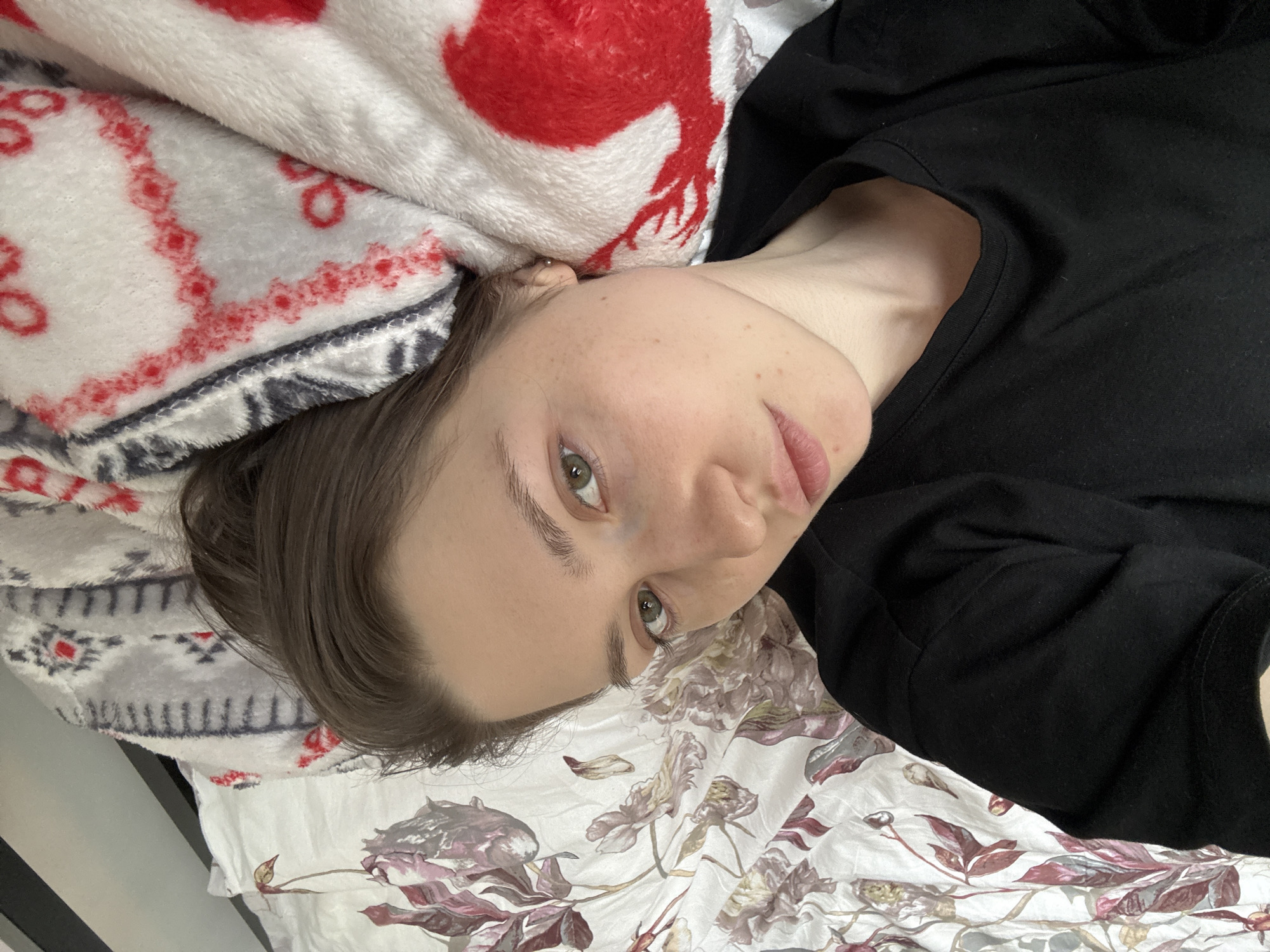 The image shows a person lying down, possibly in a bed. The person has fair skin, short brown hair, and green eyes. They are wearing a black shirt. The person is lying on a bed covered with two different patterned blankets. The top blanket has a red and white deer pattern, while the bottom blanket has a floral pattern with shades of red, purple, and brown on a white background. The lighting appears to be soft and diffused. There is no information in the image to indicate it was taken in Saint Petersburg, Russia.