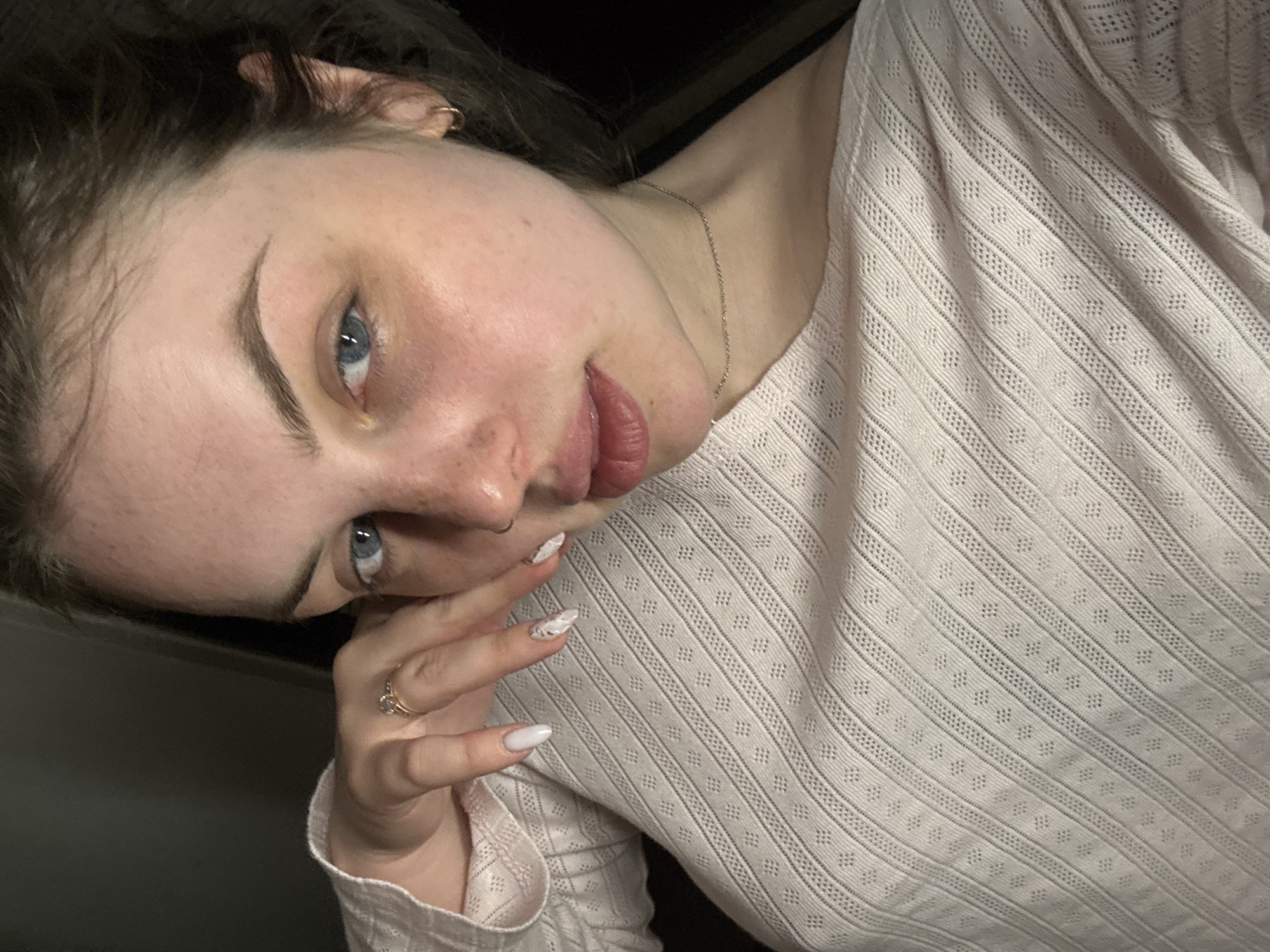 A close-up portrait features a young person, likely female, from the chest up, positioned at an angle with her head tilted. Her striking blue eyes gaze directly at the camera. She has fair skin, dark hair, and subtly defined eyebrows, with a nose piercing visible. Her lips are slightly parted, and her expression is neutral yet engaging.
She wears a light-colored, possibly light pink or beige, long-sleeved top characterized by a detailed, textured weave. The fabric displays alternating vertical bands of fine stripes and small diamond-shaped eyelet patterns. A delicate silver chain necklace is visible around her neck.
Her left hand is gently placed near her face, with manicured nails, predominantly in a light, solid color, while one nail features an intricate white design. A ring with a clear stone is worn on her left ring finger.
The background is dark and out of focus, indicating an indoor setting. The lighting is soft and appears to be ambient or artificial. The image gives the impression of a self-portrait. No text is visible. The location context provided is Chornomorskoe, Ukraine.