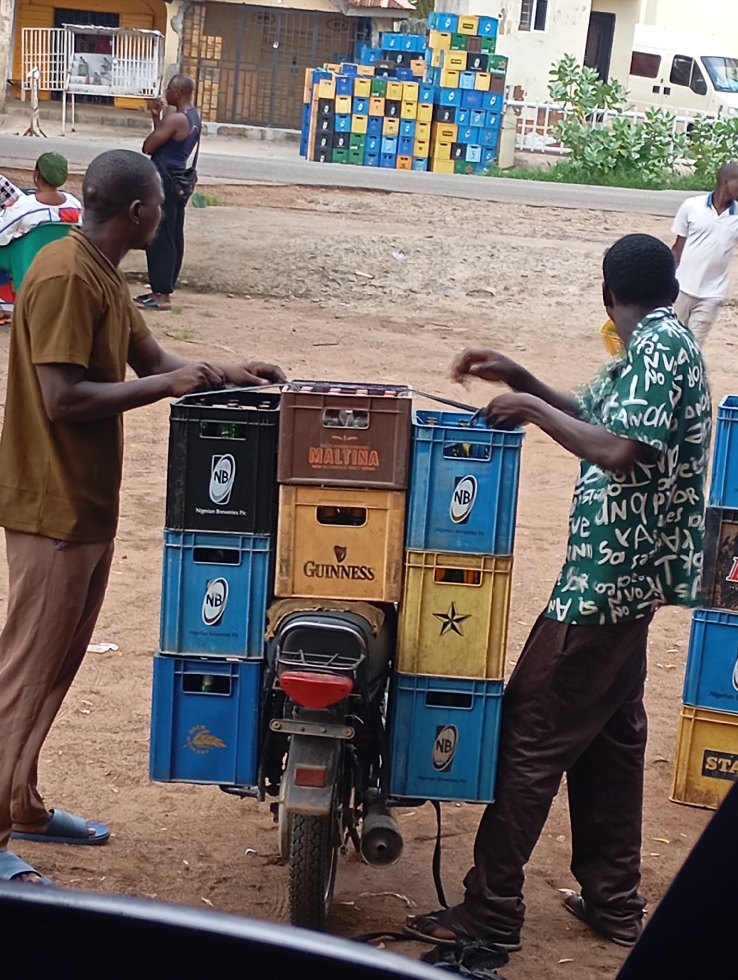 In Shendam, Nigeria, two men are seen near a motorcycle loaded with crates of beverages. The man on the left, wearing a brown t-shirt and brown pants, stands next to the motorcycle, his hands resting on top of the crates. The man on the right, in a green patterned shirt and dark pants, appears to be adjusting the load. The motorcycle itself is positioned in a dirt area, with several stacks of beverage crates visible behind and to the side of it. In the background, buildings and more stacks of crates suggest a commercial or market area. A white van is parked in the background. The scene suggests a busy commercial environment, possibly a distribution point for beverages.