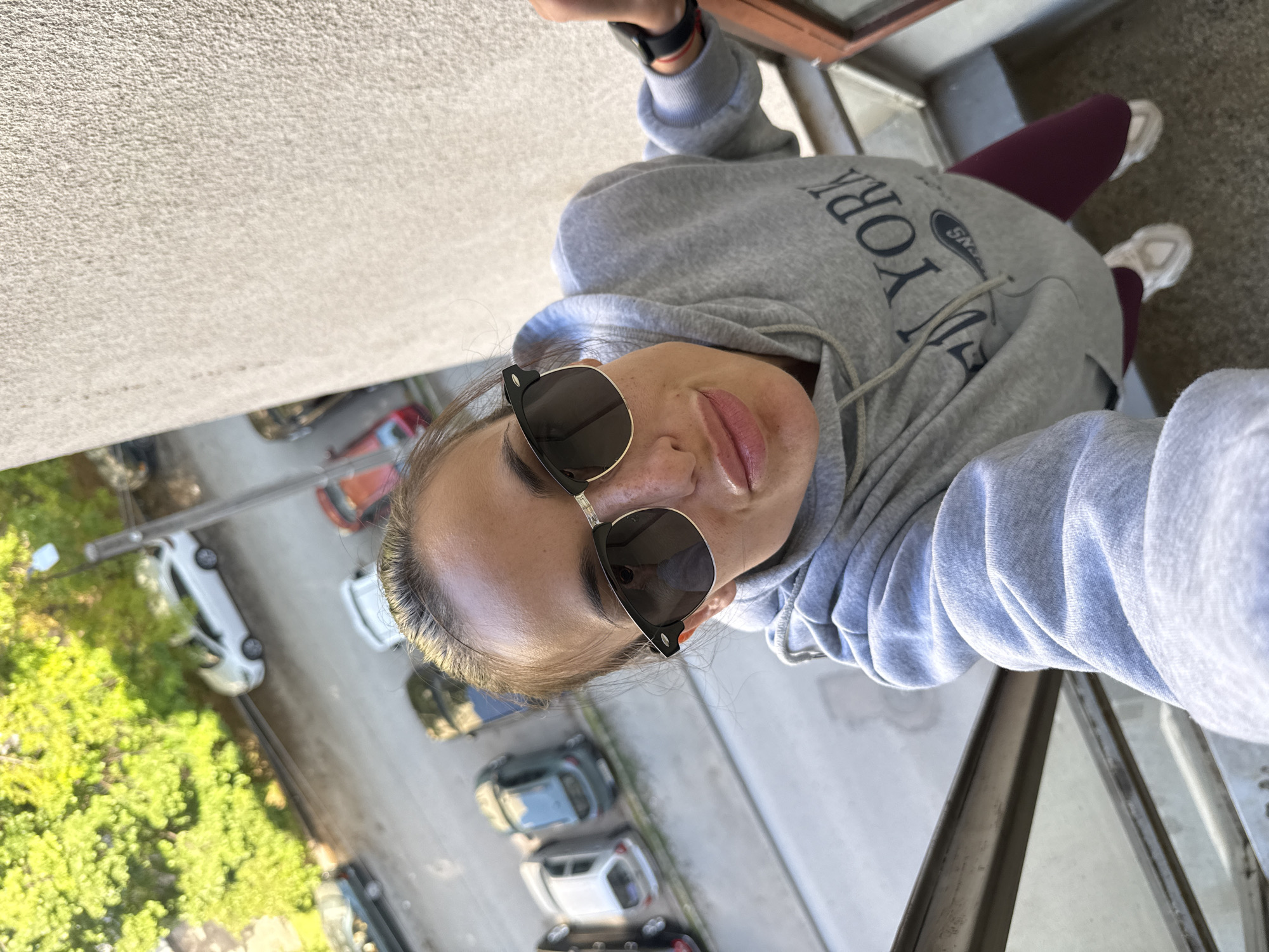 A woman is taking a selfie from a high angle, possibly a balcony. She's wearing sunglasses and a gray hoodie with "New York" printed on it, along with maroon leggings and white sneakers. Below, a street features several parked cars (white, gray, black, red) and green trees. Based on the car license plates, the picture was taken in Bulgaria, and based on the architecture of the buildings and the city's overall environment, it is highly likely that the location is in Pleven, Bulgaria. The woman is wearing a fitness tracker on her wrist.