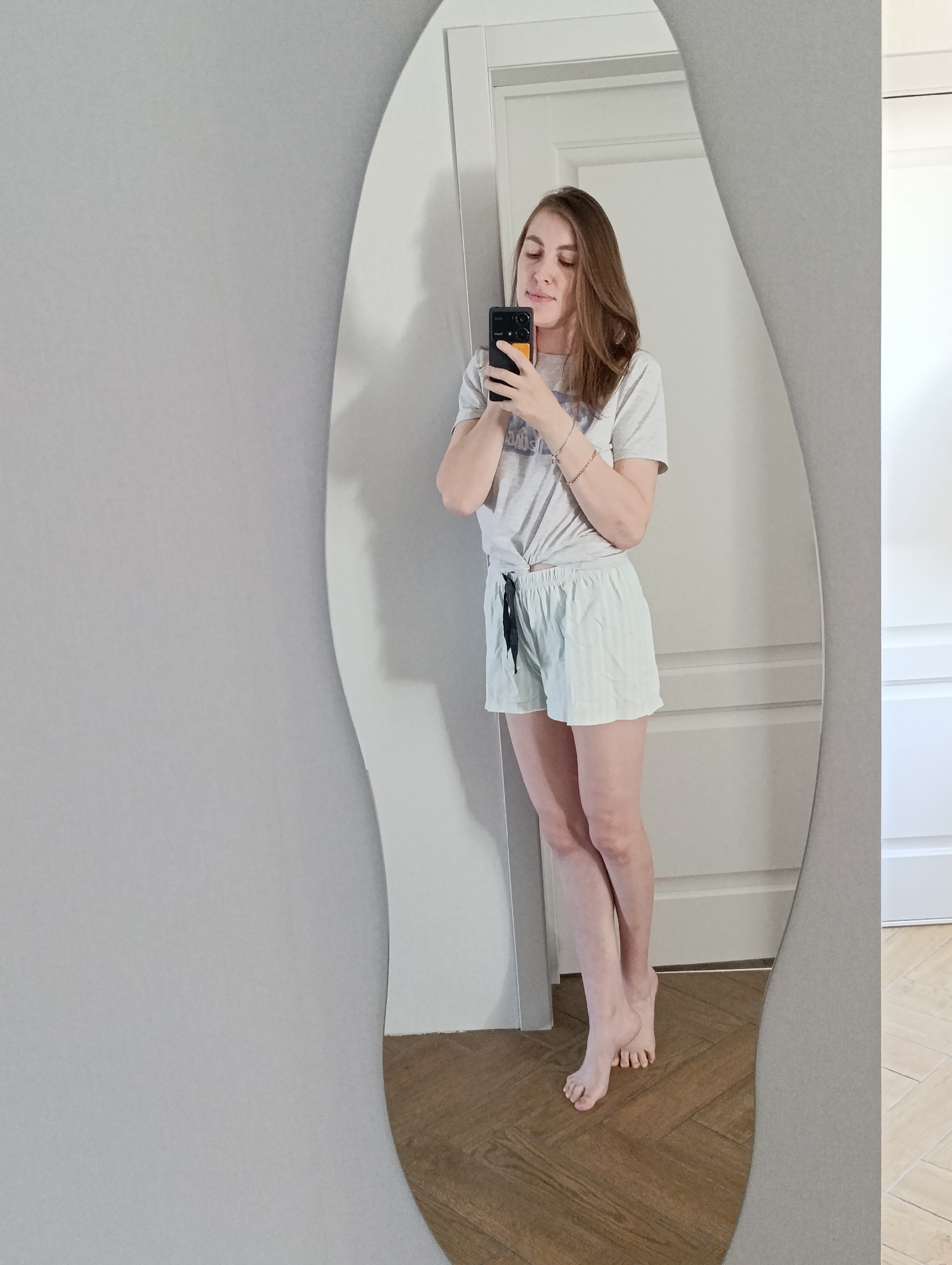 A woman is taking a selfie in a full-length mirror. She is fair-skinned with brown hair. She is wearing a gray t-shirt tied in a knot and light blue striped shorts with black ties. She is barefoot. She is holding a black and yellow smartphone. The background includes a white wall, a white door with panels, and a herringbone pattern wood floor. The mirror has an irregular, wavy edge. The location appears to be indoors. The presence of a herringbone wood floor and a paneled door suggests a modern or renovated interior, though determining the exact location in Chaikovsky, Russia, from the image alone is impossible.