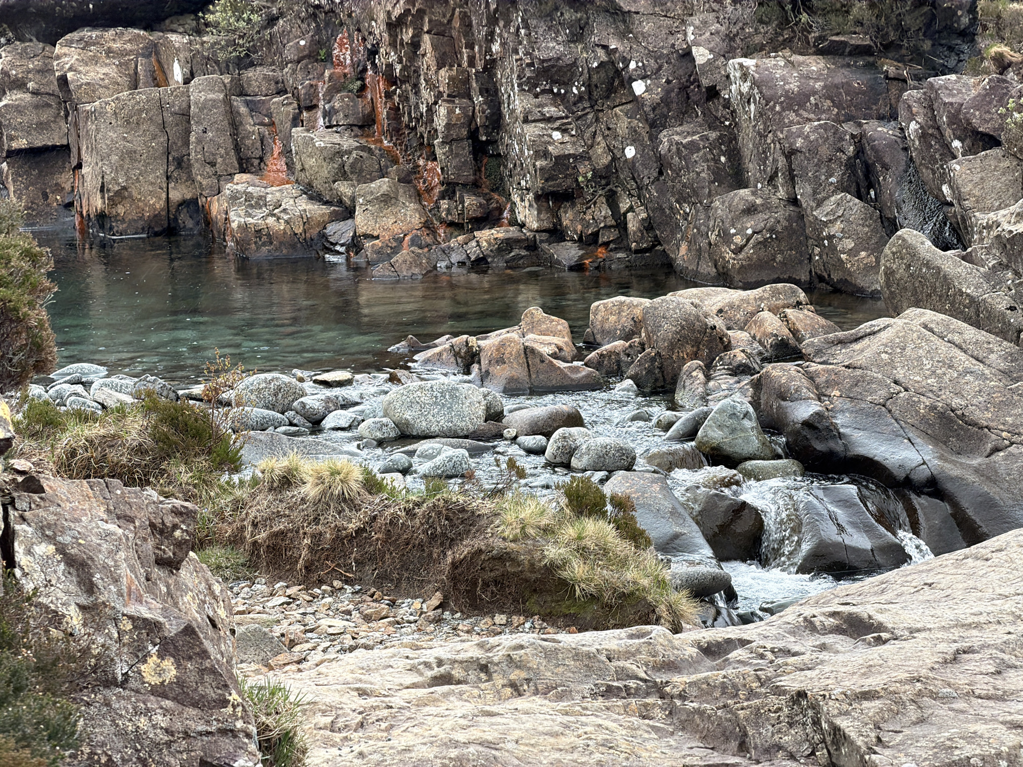 The image depicts a rocky landscape with a small stream flowing through it, likely located in the Isle of Skye, United Kingdom. The foreground is dominated by large, light-colored rocks, textured with cracks and small patches of vegetation. A section of the stream is visible, containing smooth, gray rocks of various sizes. Patches of coarse grass and exposed soil appear along the stream's edge. In the background, darker, more rugged rocks form a wall beside a deeper, still pool of water. Some of these rocks show streaks of reddish-brown coloration. The water in both the stream and the pool appears clear. No people are visible in the image.