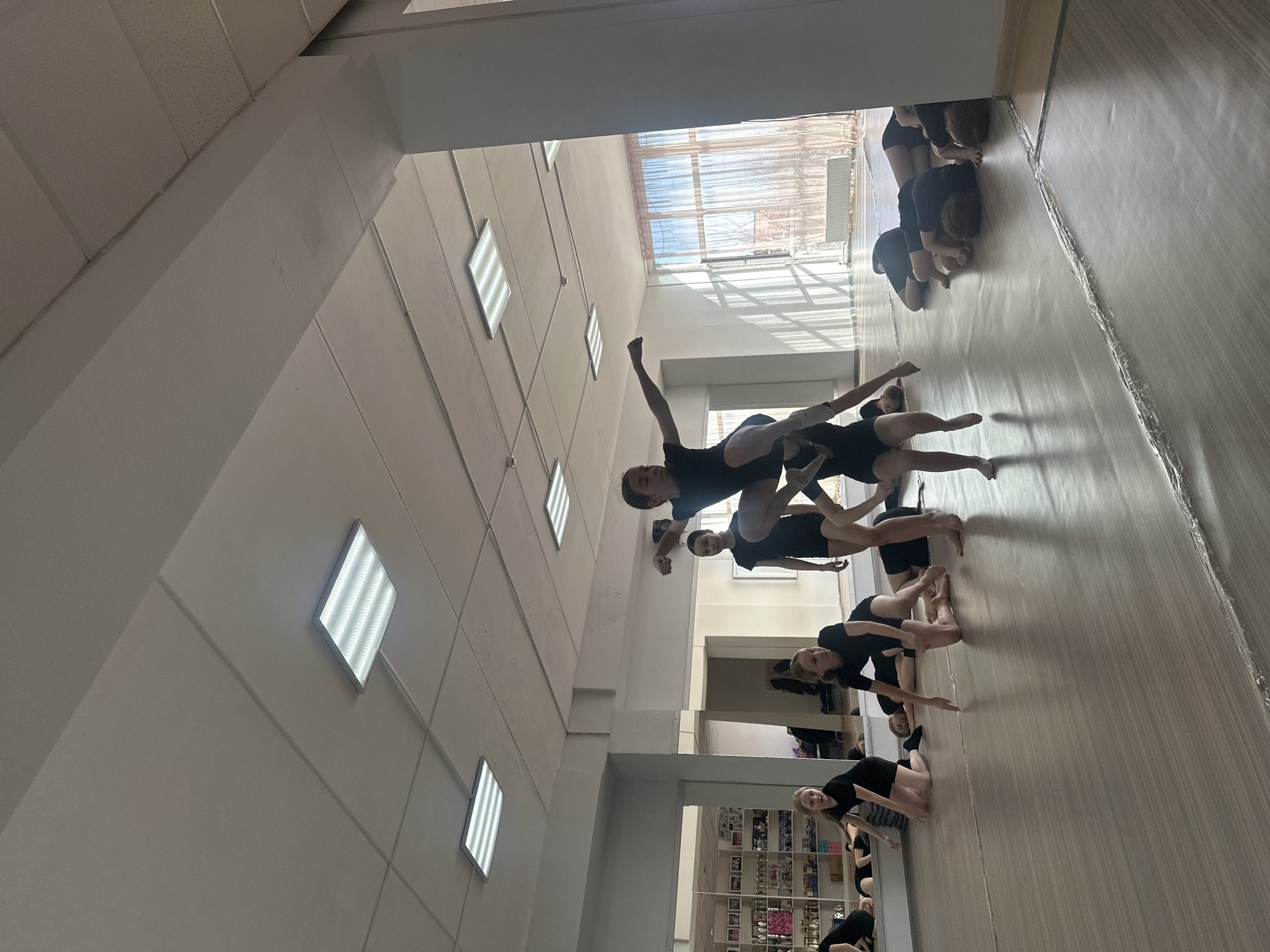 The image shows a group of dancers in a studio. The studio has a hardwood floor, white walls, and fluorescent lighting. A large window is visible in the background. The dancers are wearing black leotards and shorts. They are arranged in lines performing various dance moves. The location is identified as Votkinsk, Russia.
