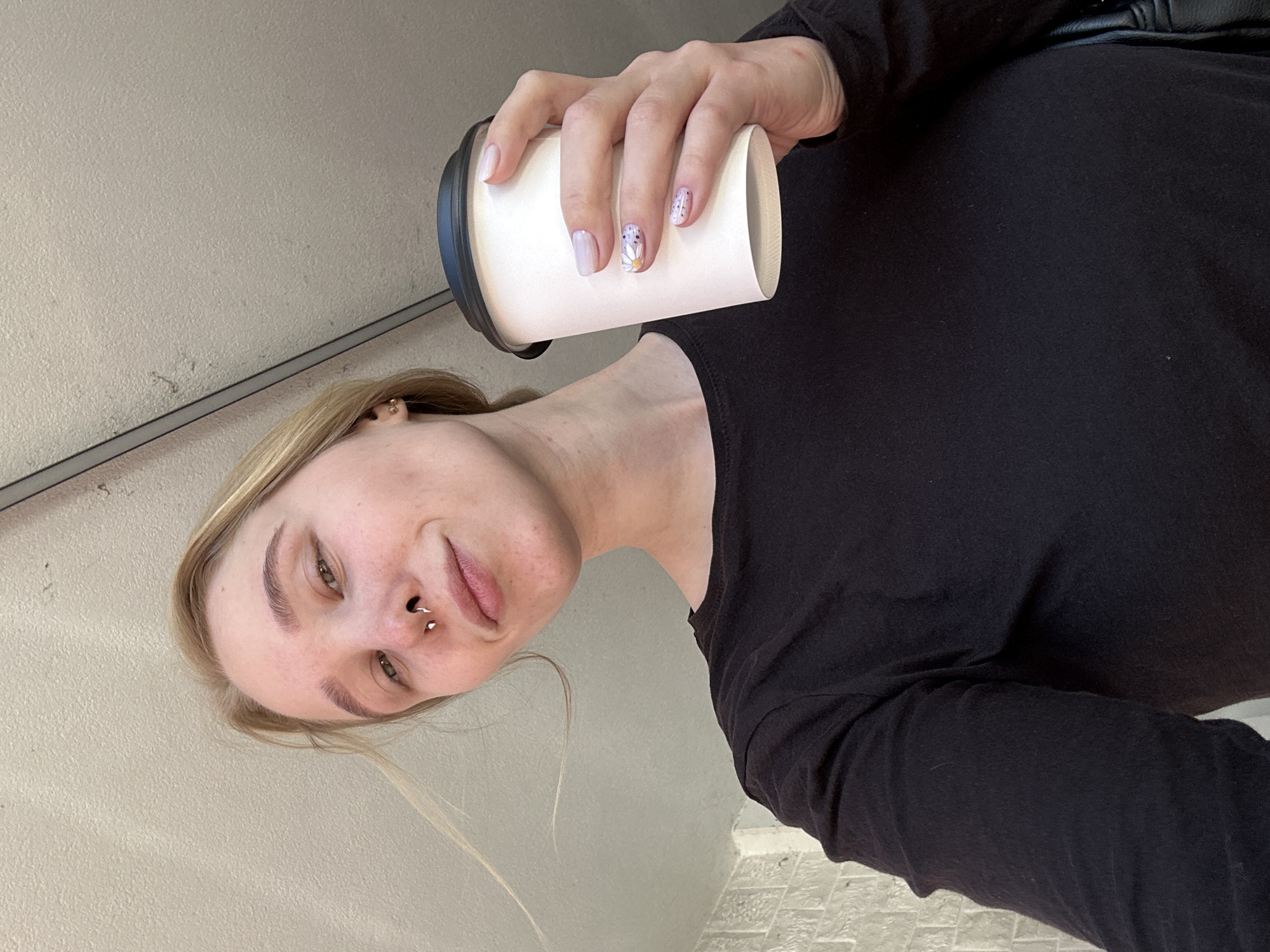 The image is a rotated, low-angle selfie of a young person, likely taken in Budva, Montenegro. The individual, who appears female, has blonde hair and fair skin, and looks directly into the camera with a neutral to slightly smirking expression. She has a septum piercing and small stud earrings in both earlobes. Her right hand, featuring manicured nails with light purple polish and designs (a daisy on the ring finger and dots on the pinky), holds a white disposable coffee cup with a black lid. She is wearing a black long-sleeved top. The background consists of a plain, textured, light gray or off-white wall or ceiling, with a dark horizontal line running across it. The lighting is bright and even, suggesting it is daytime. There is no visible text in the image.