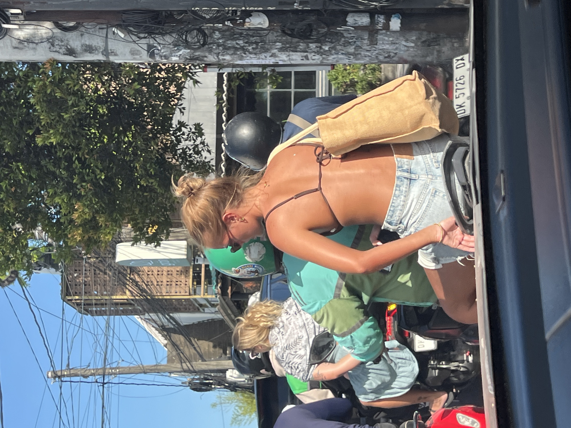 The image depicts a scene from a street in Unknown, Unknown. A woman with blonde hair pulled back in a bun, wearing a brown bikini top and denim shorts, is riding on the back of a scooter. She is carrying a tan-colored bag. The scooter is driven by a person wearing a green helmet and a light blue shirt. Behind them, another scooter carries a person wearing a black helmet, a white patterned shirt, and light blue shorts. The license plate on the back of the first scooter reads "DK 5726 DX". Overhead, a tangled web of electrical wires is visible against a backdrop of buildings and lush green trees. The image is taken from inside a vehicle, capturing the scene through the window frame on the right side of the frame.