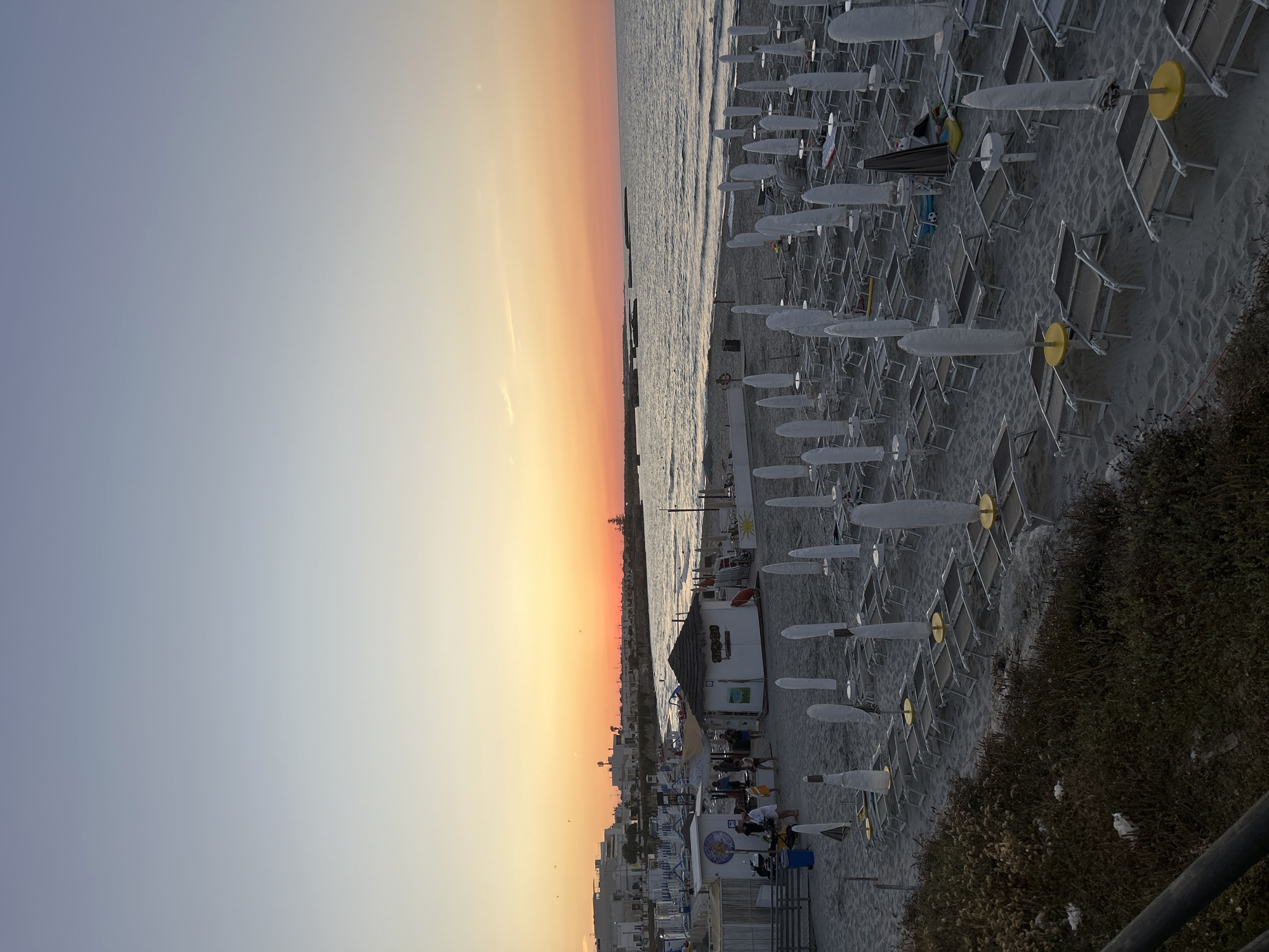 An overhead, high-angle, wide shot captures a beach scene at dusk in San Foca, Italy. The sky transitions from a pale blue at the top to a vibrant orange and pink gradient at the horizon, with the sea gently lapping the shore. Numerous rows of empty lounge chairs and closed umbrellas are arranged on the sandy beach. A few small, white buildings with thatched roofs are visible on the sand, with a few people walking around them. To the left, a low wall and dark foliage line the foreground of the image, with a wooden railing partially visible at the bottom left corner. The overall atmosphere is serene, with the setting sun casting a warm glow over the tranquil beach.