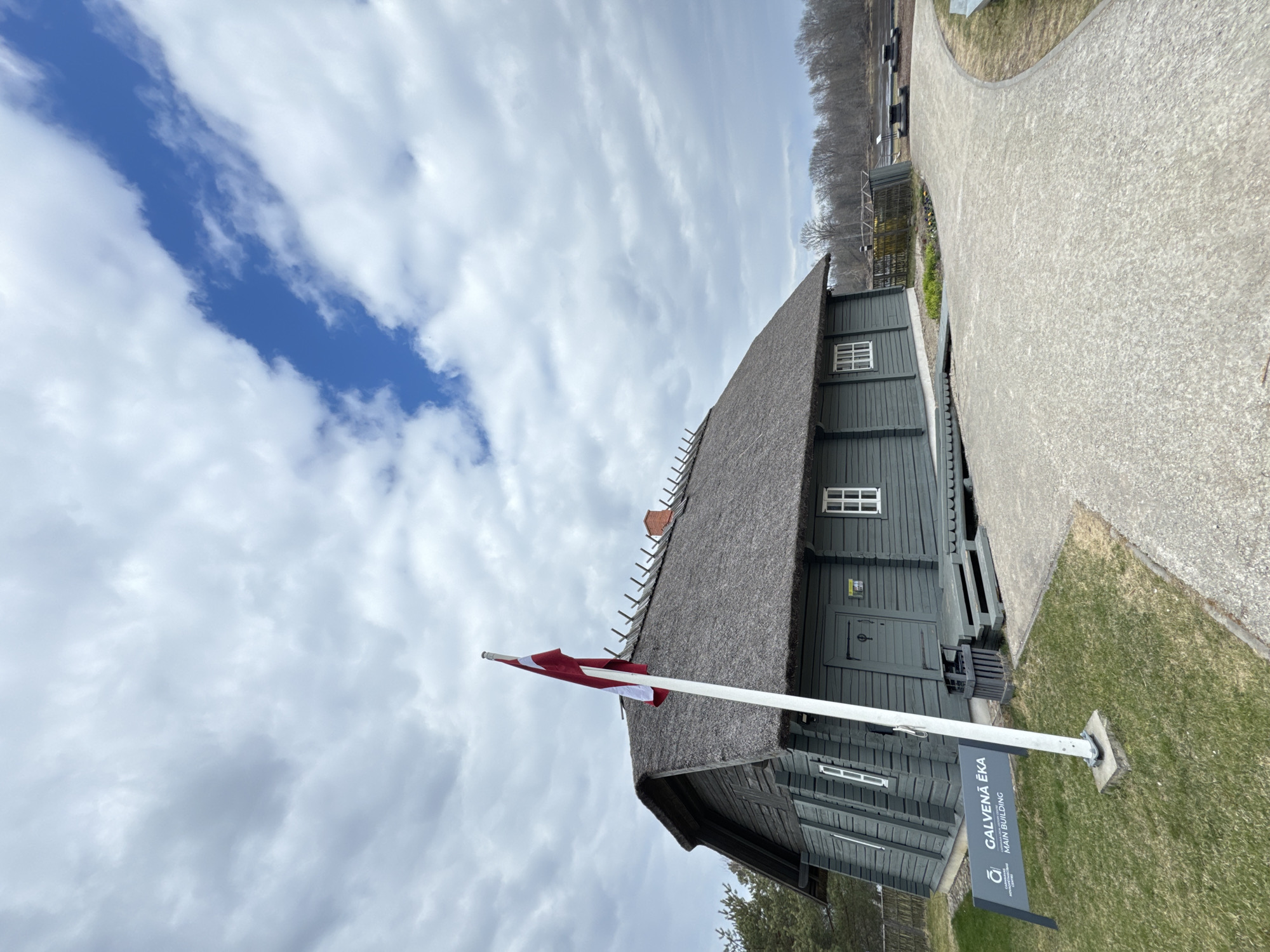 The image depicts a rural scene in Carnikava, Latvia. A traditional wooden building with a thatched roof is the main subject. A Latvian flag stands in front of the building, and a sign reads "Galvenā ēka" (Main Building). The surroundings include a gravel path, grassy areas, and trees in the background. The sky is overcast with scattered clouds.