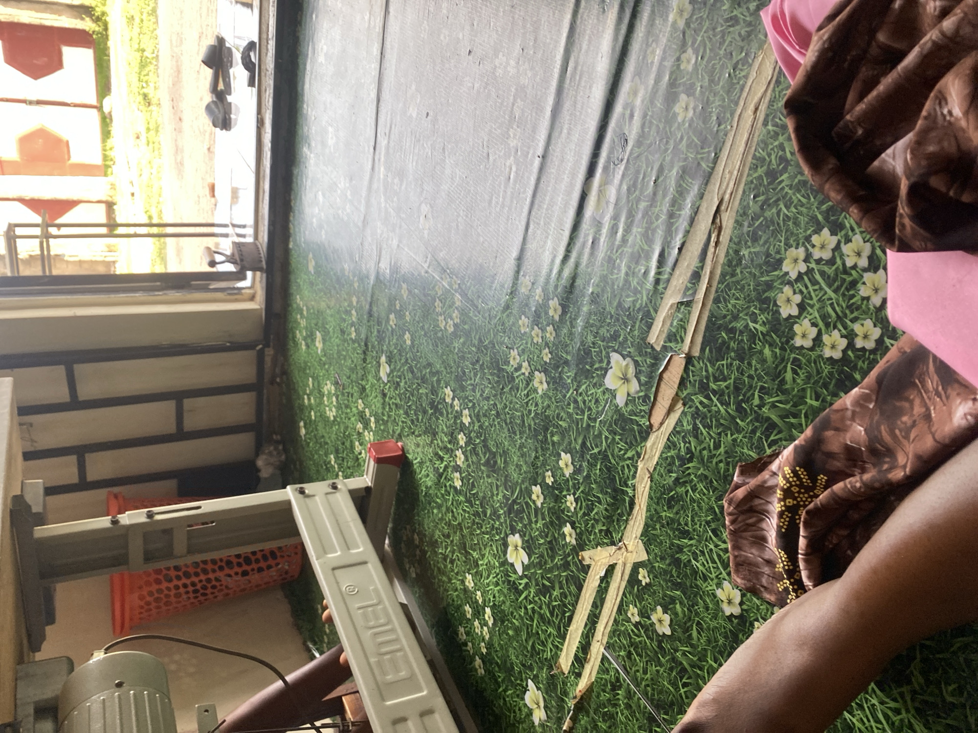 The image depicts the interior of a room in Jauro Yinu, Nigeria, likely a workshop or storage area. The floor is covered with a green, grassy print with white flowers. A sewing machine with a light-colored motor is visible on the left, partially obscured by a brown tubular object. An orange plastic basket is positioned behind the sewing machine. In the foreground, a person's legs and arms are visible, draped with brown fabric featuring yellow patterns and a pink garment. A section of the floor in the middle ground is covered by a dark, reflective material, possibly a mat or tarp. Further back, an open doorway reveals an outdoor scene with sunlight, green vegetation, and what appear to be red and white hexagonal structures, possibly part of a fence or building. A rusty iron is placed on the tiled floor near the doorway, and several black sandals are scattered near the entrance.