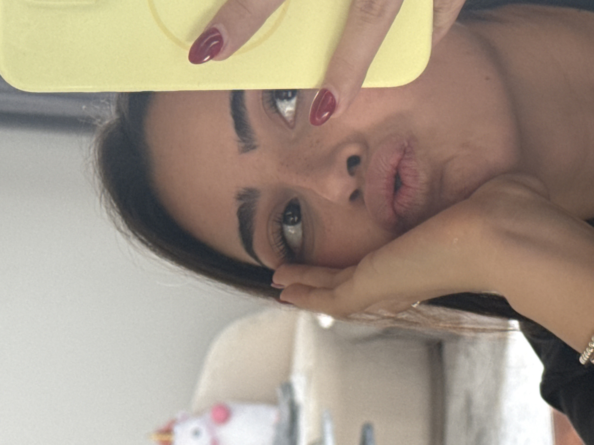A young woman is taking a selfie with a yellow phone case. She is making a duck face and holding her hand to her cheek. The background is out of focus, but appears to be an indoor setting with furniture and a blurry unicorn toy. The woman has long dark hair and is wearing a black shirt. Her nails are painted a dark red. The image is taken in the city of San Bernardino Tlaxcalancingo, Mexico.